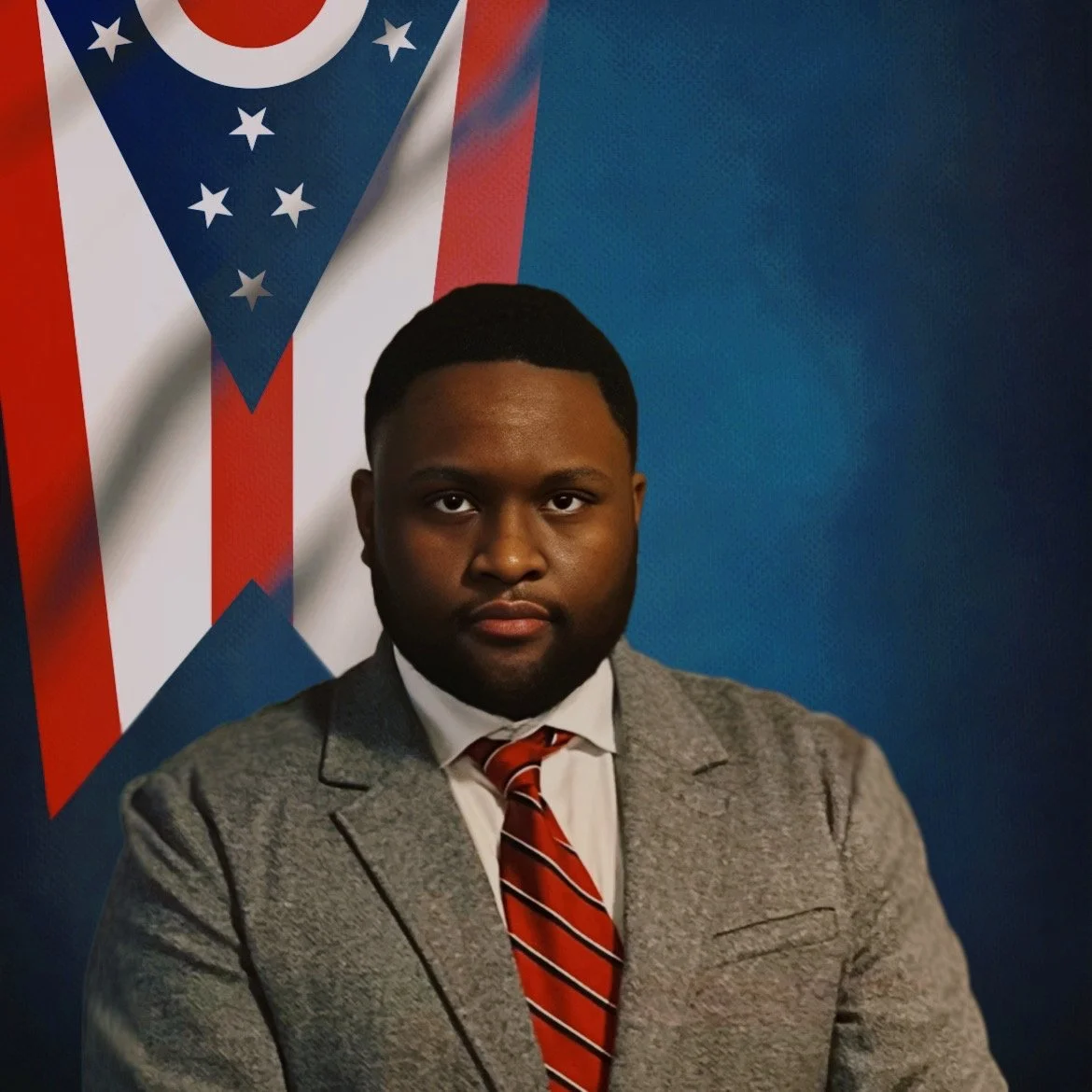A man in a gray suit, white shirt, and red striped tie, standing in front of a blue background with the Ohio state flag displayed prominently behind him.