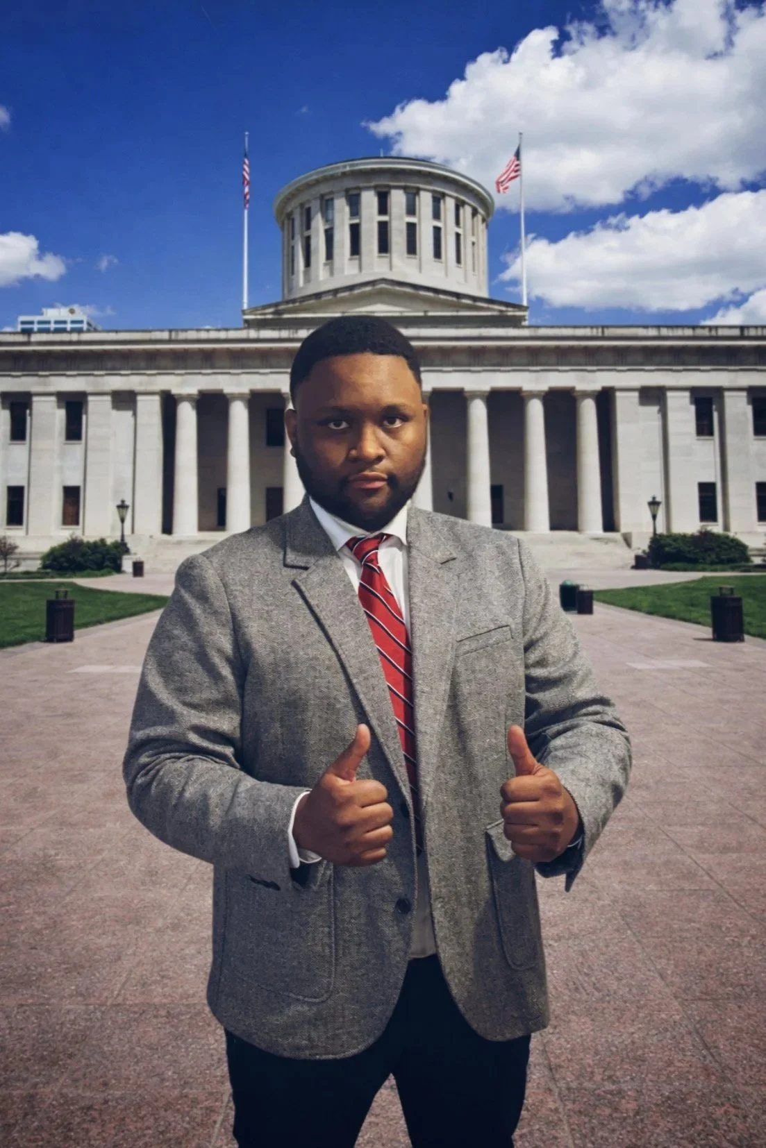 A man wearing a grey blazer, white shirt, and red tie giving two thumbs up in front of a government building with columns and a dome, under a partly cloudy sky.