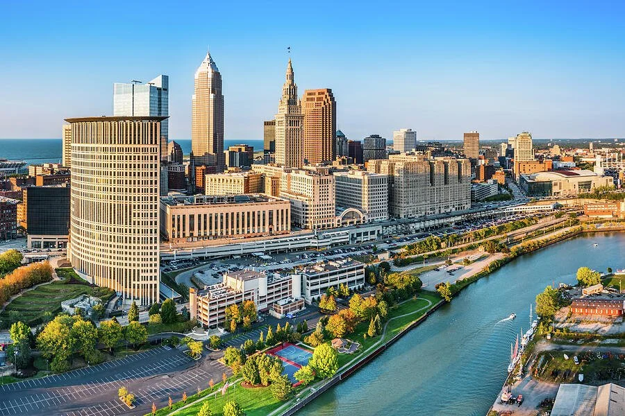 A city skyline with tall skyscrapers next to a river, with parks and parking lots in the foreground.