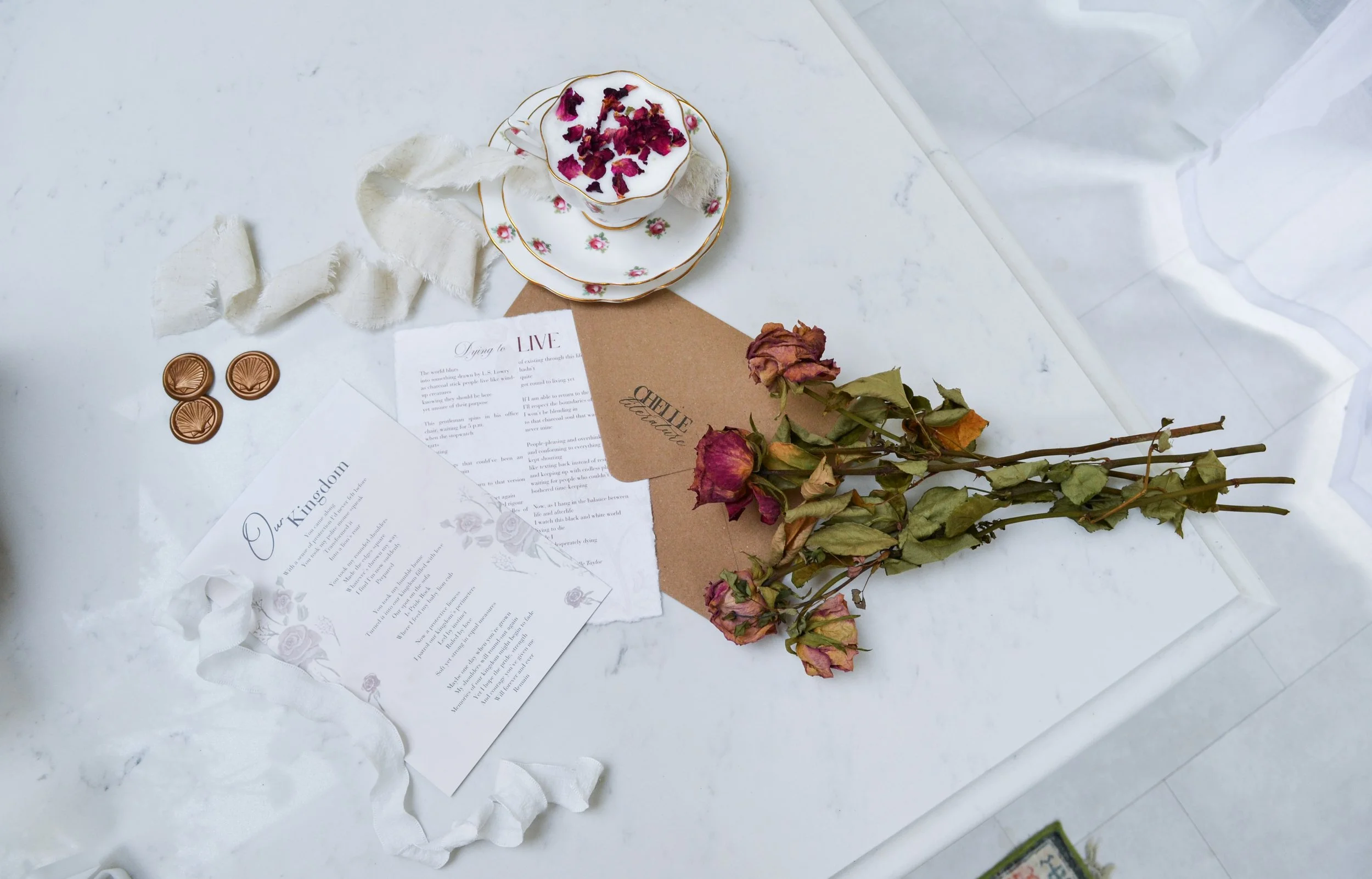 A white marble table with dried pink roses on a brown paper, a white and gold tea cup with petals inside, a matching saucer and plate, three round chocolates, and two notes with handwritten and printed text.