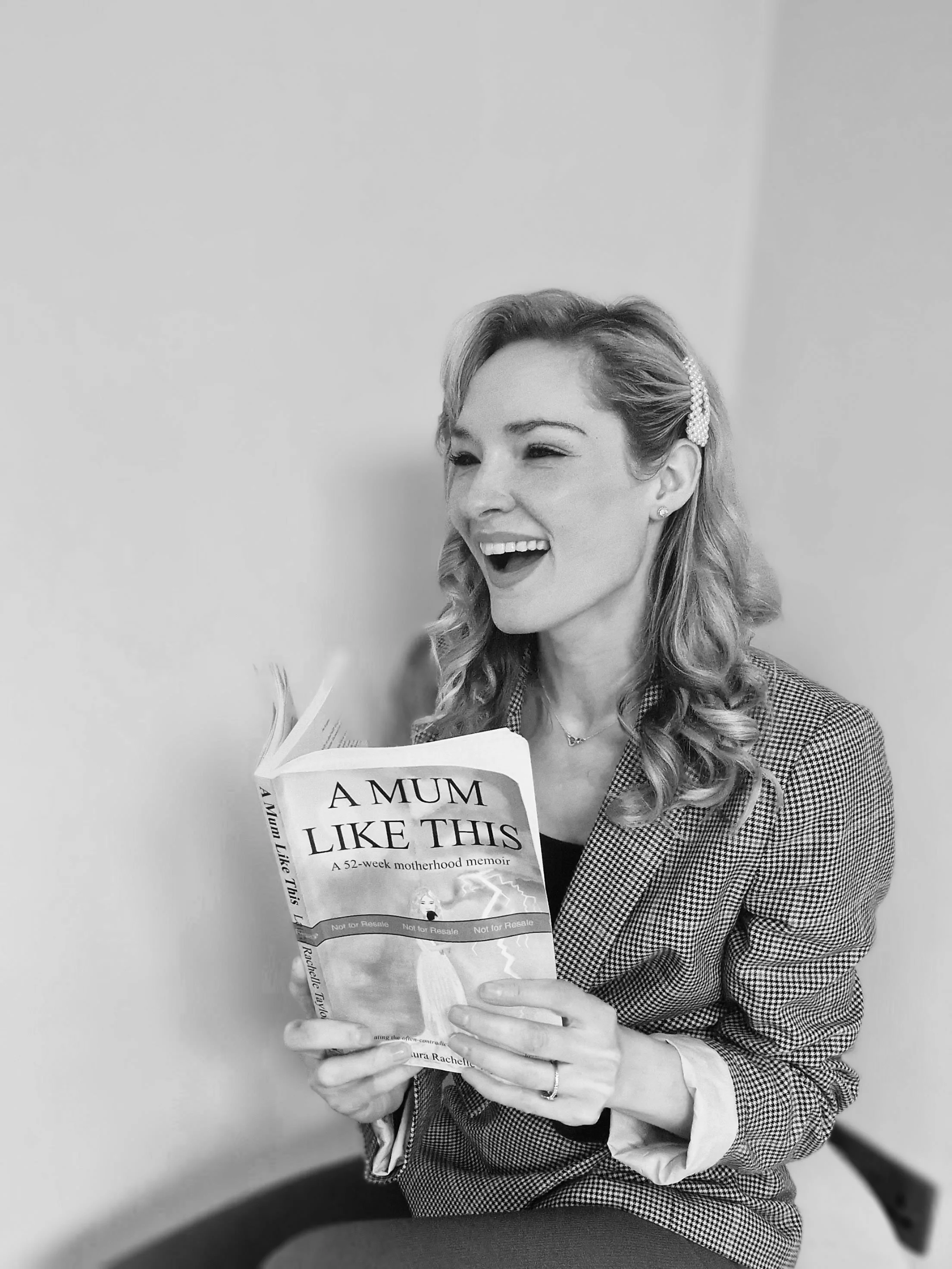 A woman with wavy hair wearing a checkered blazer and earrings is sitting on a chair, smiling while holding and looking at a book titled 'A Mum Like This'.