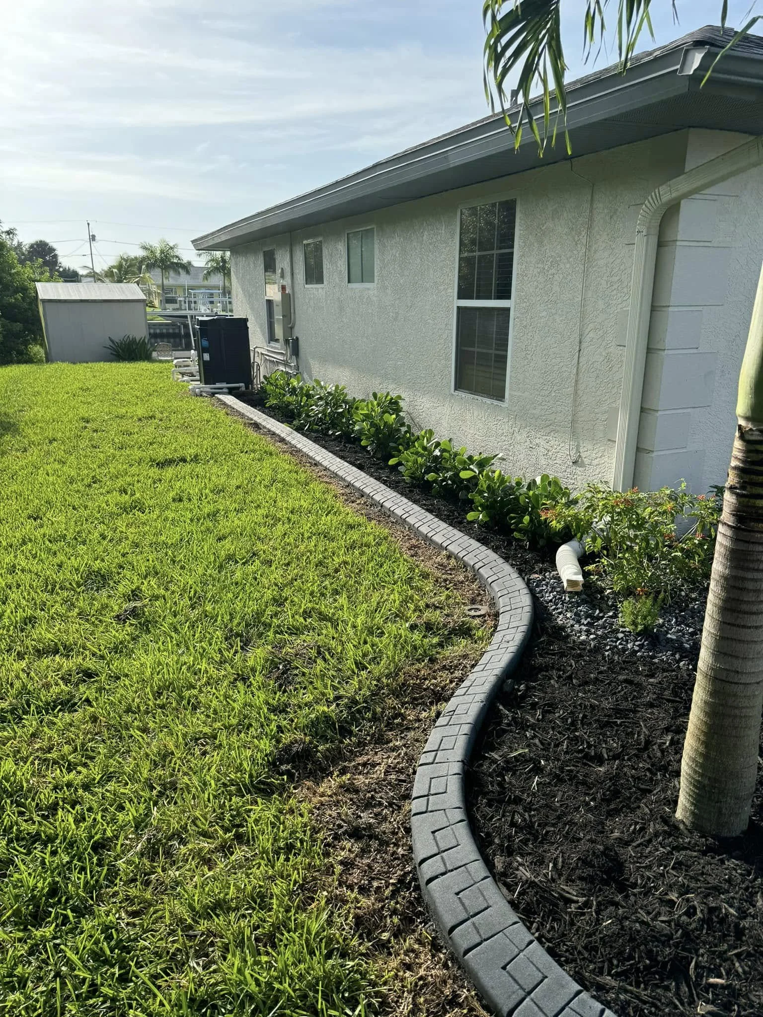 A backyard with a green lawn, a garden bed with shrubs and plants along the side of a white house, and a curved plastic edging separating the grass from the garden.