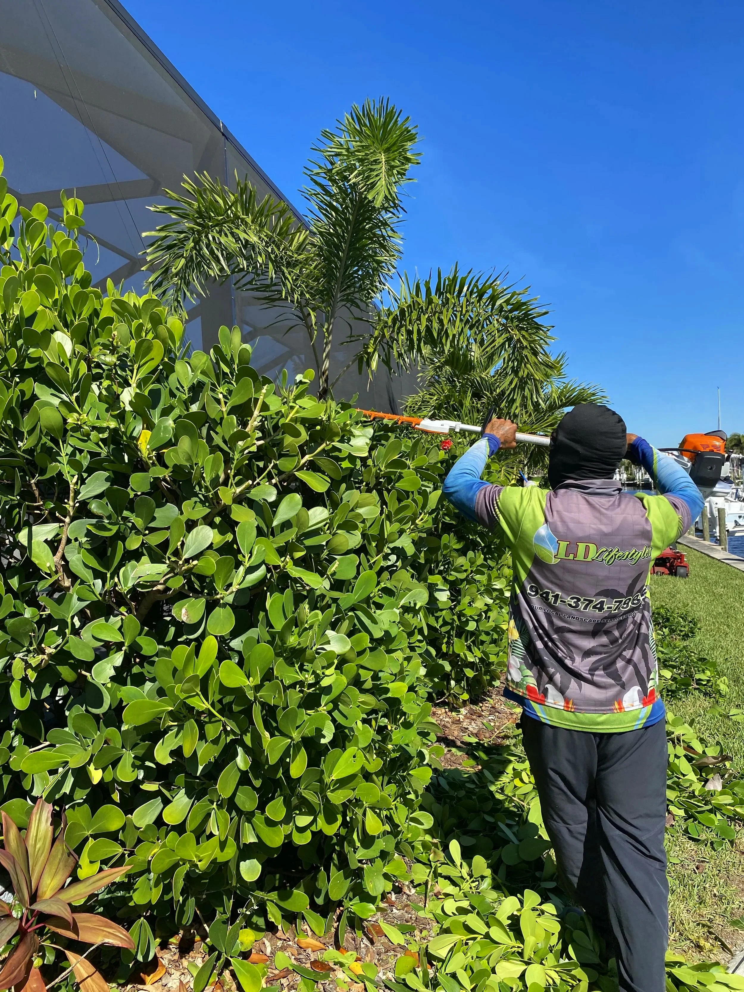 LD Lifestyles worker trimming bushes with a pole hedge trimmer in a landscaped area near Punta Gorda marina.