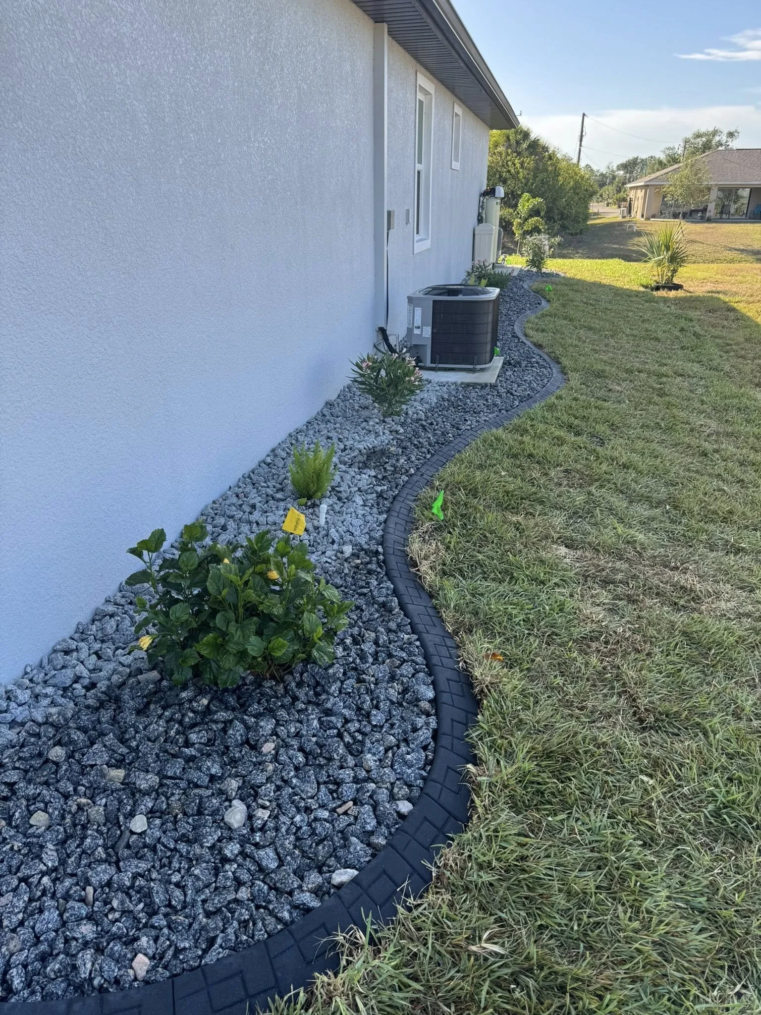 A landscaped backyard with a white exterior wall, black curbing, and decorative rocks, featuring some plants near the house and a grassy area.
