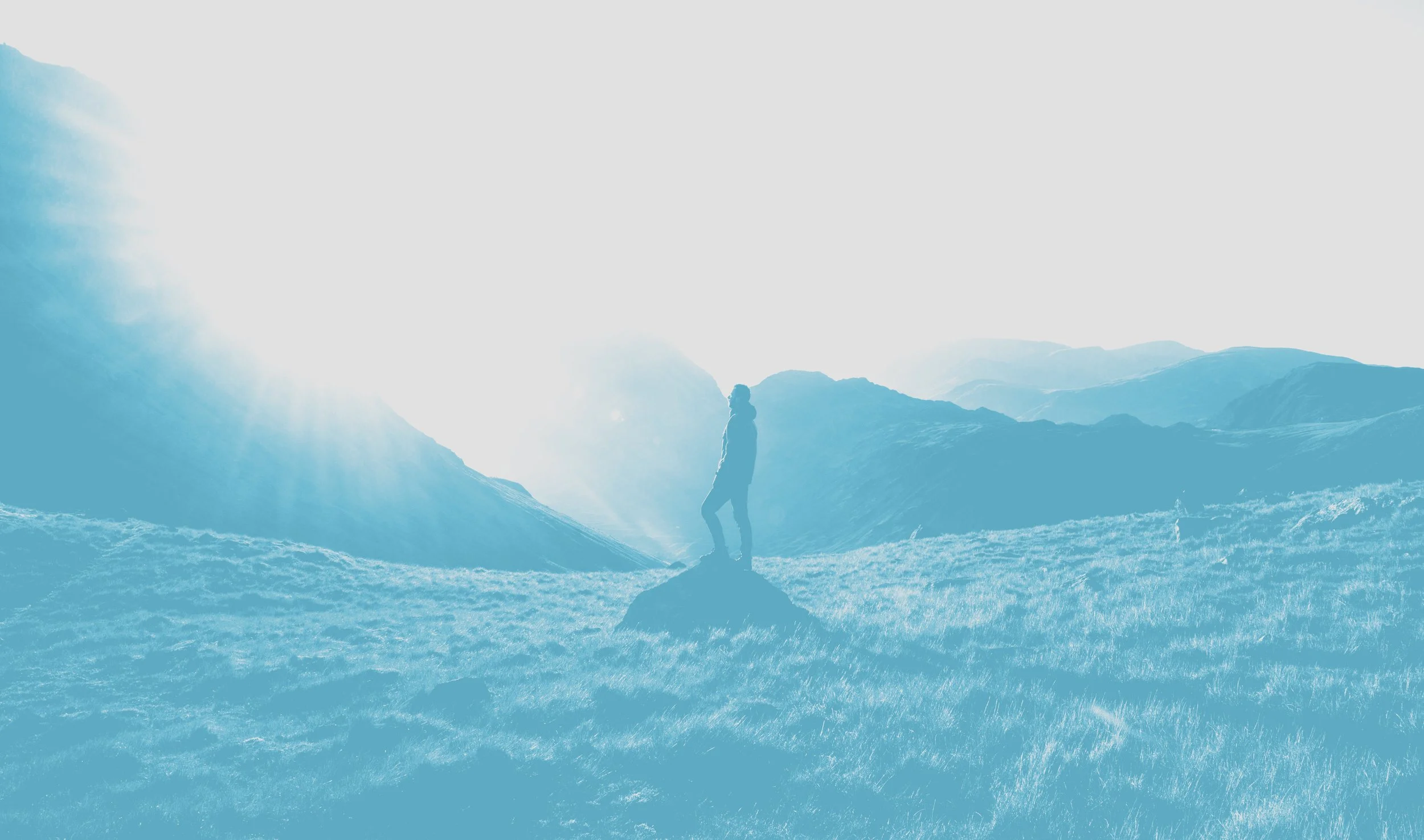 A person standing on a rock in a grassy landscape with mountains in the distance and the sun shining brightly.