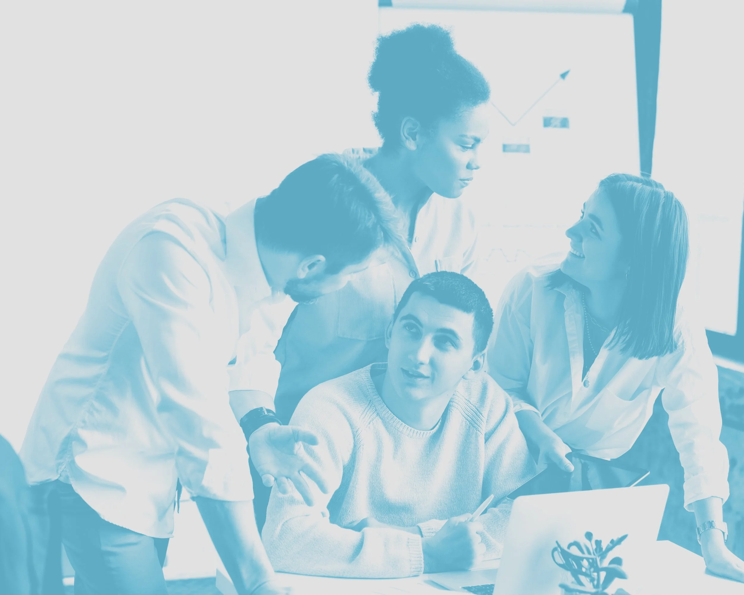 Group of five diverse professionals in a business meeting, with one person sitting and four standing around, engaging in discussion, in a conference room with a presentation chart in the background.