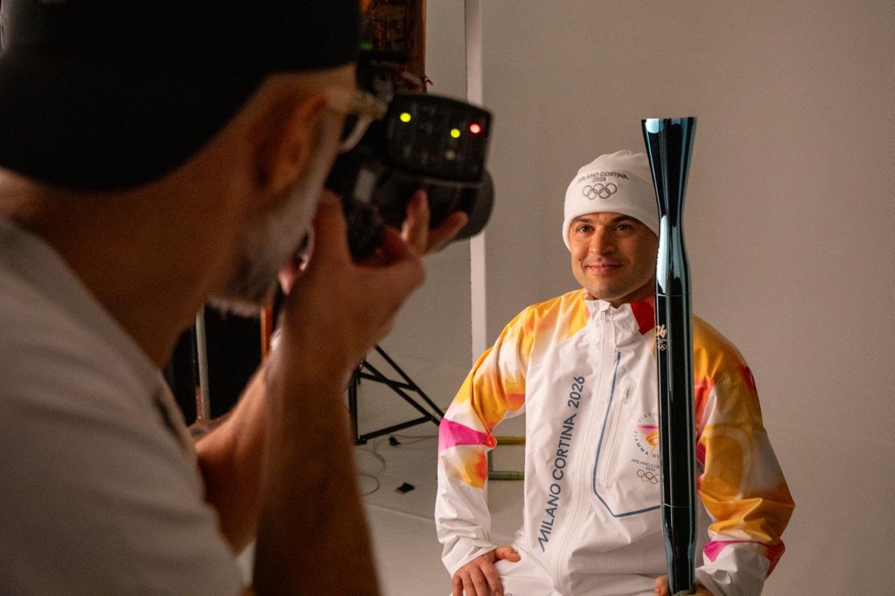 Atleta olimpico sorridente in abbigliamento invernale con sci, posa per la foto mentre un fotografo lo scatta, sfondo neutro.