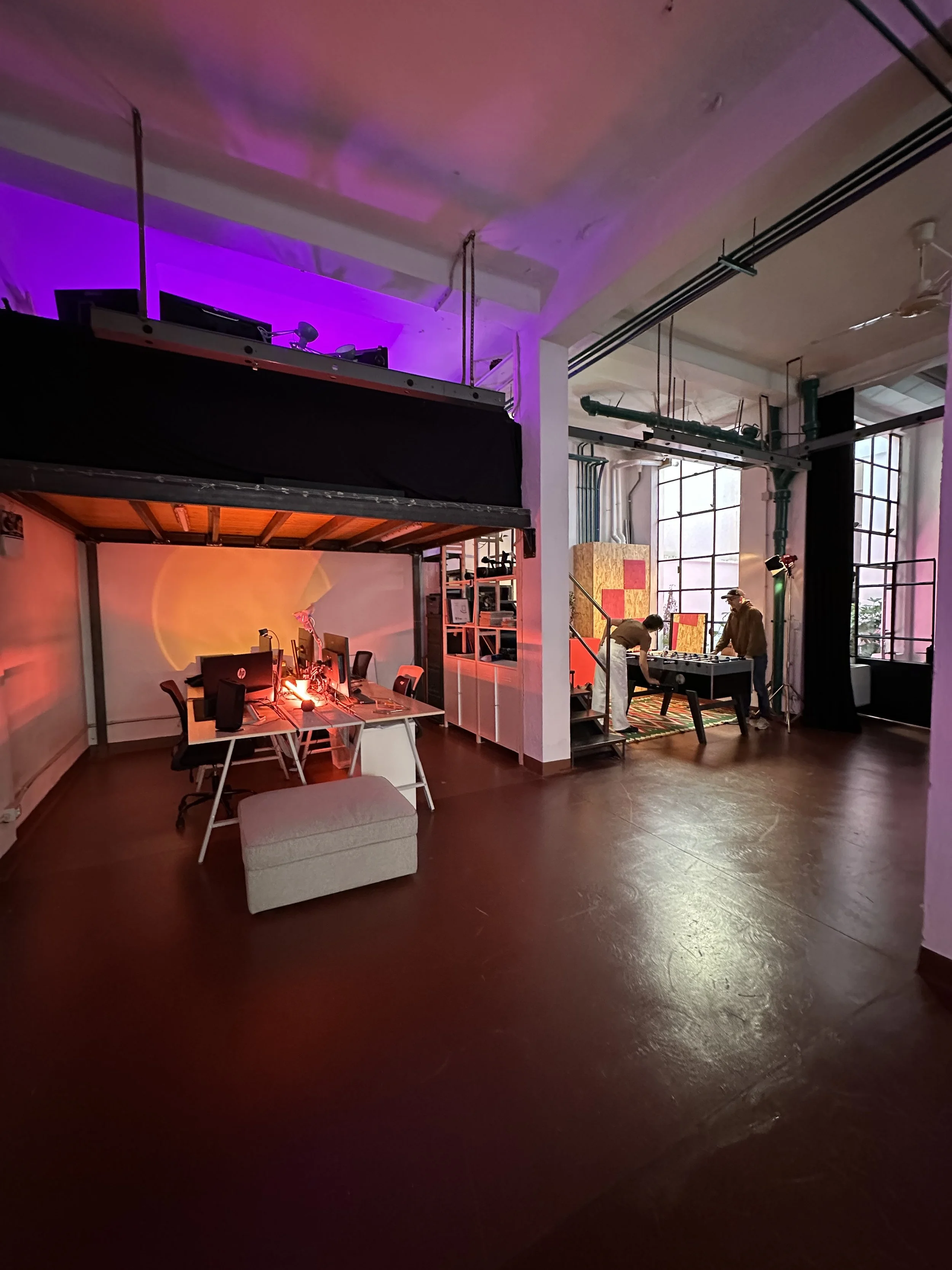 Interno di un loft con pareti di grandi finestroni. Spazio aperto con tavolo e computer, due persone che giocano a calcio balilla, illuminazione con luci colorate.