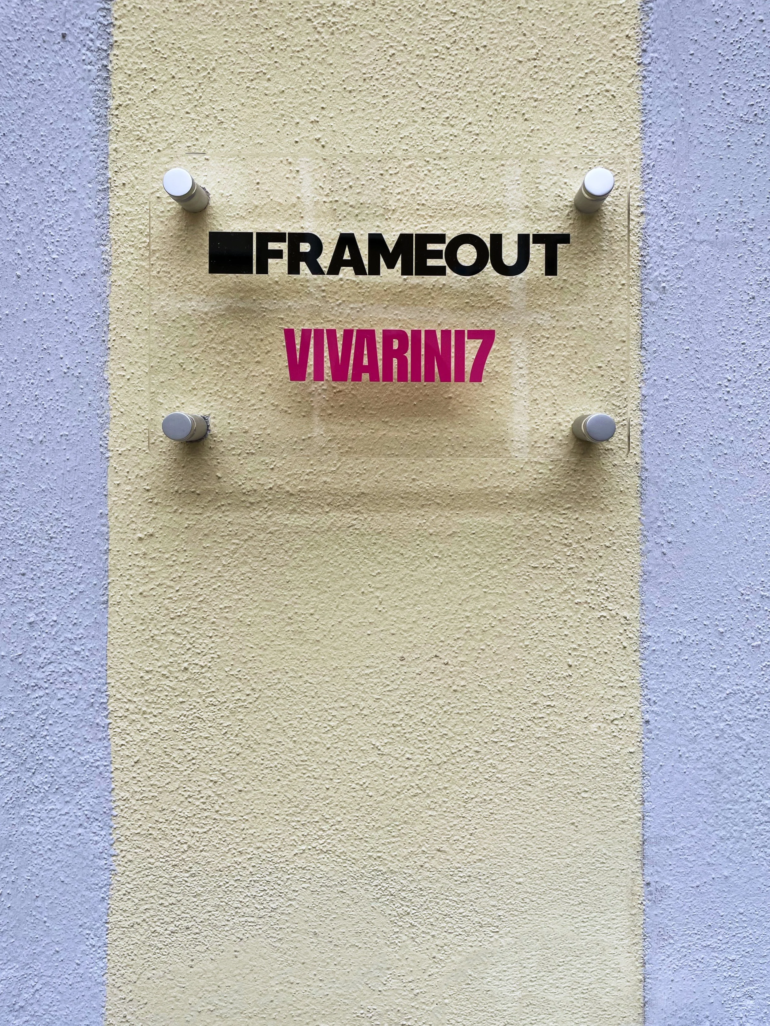 Pannello segnaletico con il logo Frameout in nero e la scritta VIVARINZ in rosa sopra uno sfondo giallo becco con pareti laterali grigie.