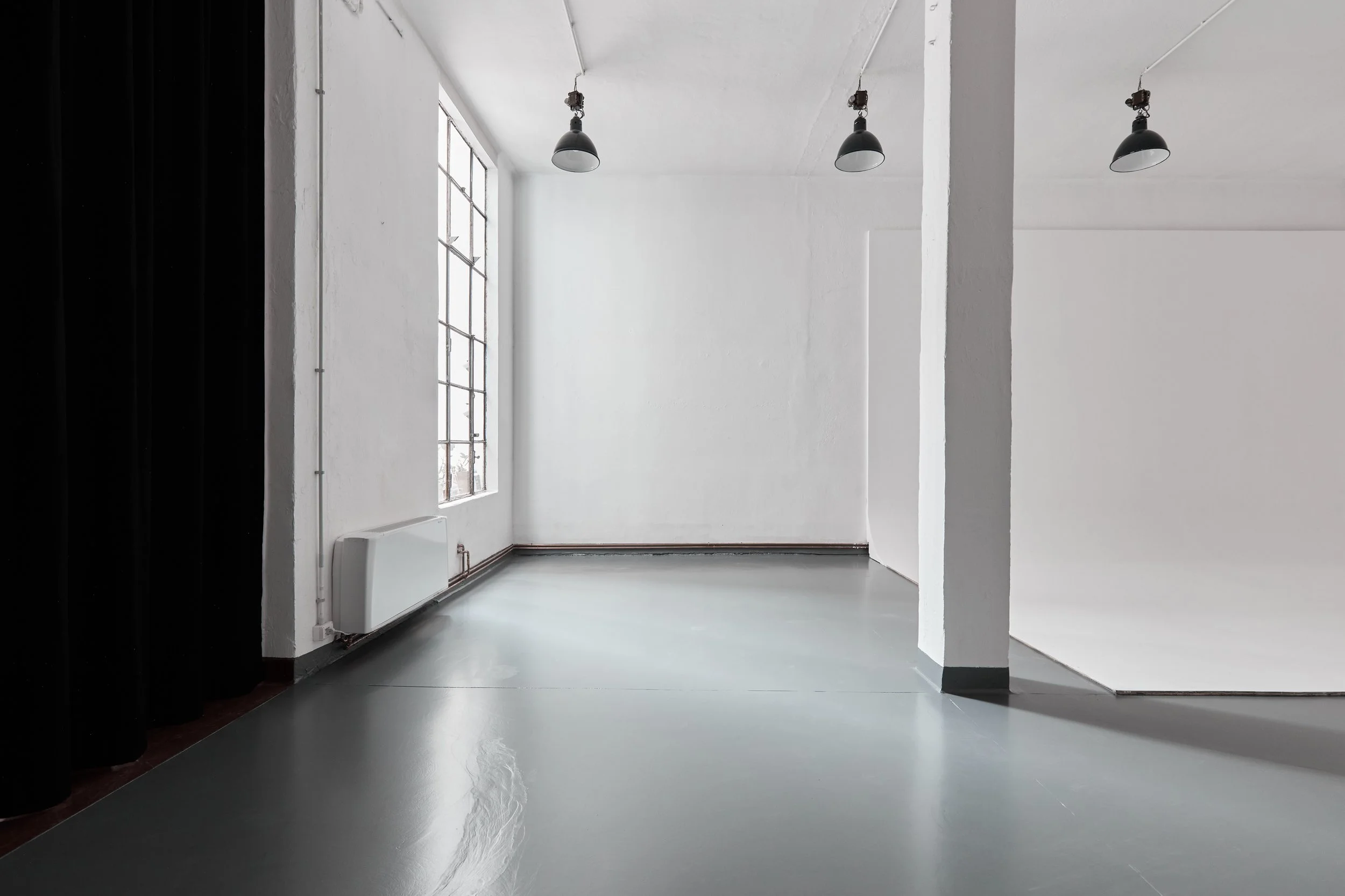 Interno di una stanza vuota con pareti bianche, grande finestra con inferriate, luci pendenti nere, pavimento lucido grigio e radiatore bianco sotto la finestra.