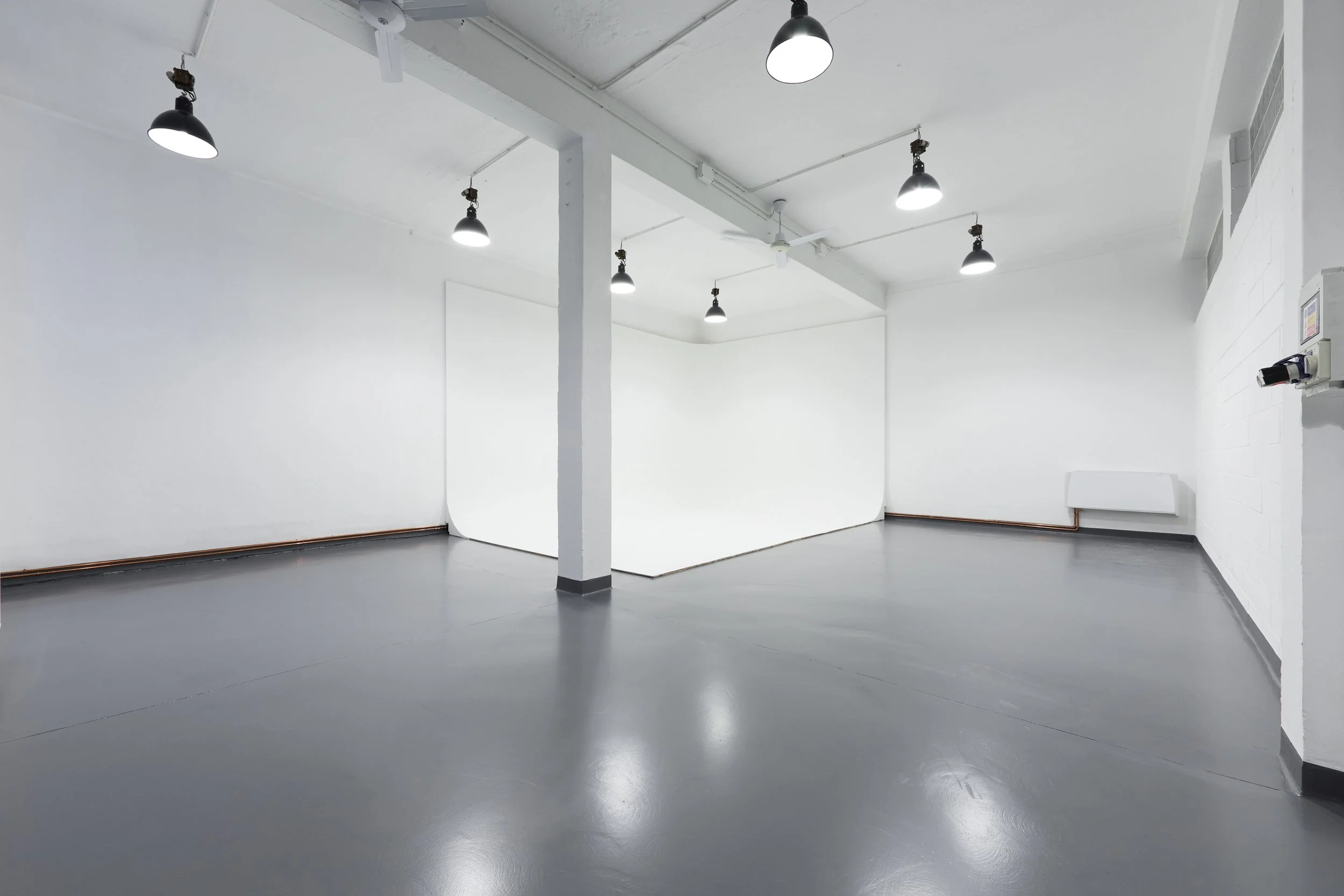 Stanza vuota con pareti bianche, pavimento grigio lucido, illuminazione da lampade a soffitto, parete curva e pareti con elementi di riscaldamento e impianto di sicurezza. Studio fotografico con ciclorama ad L.