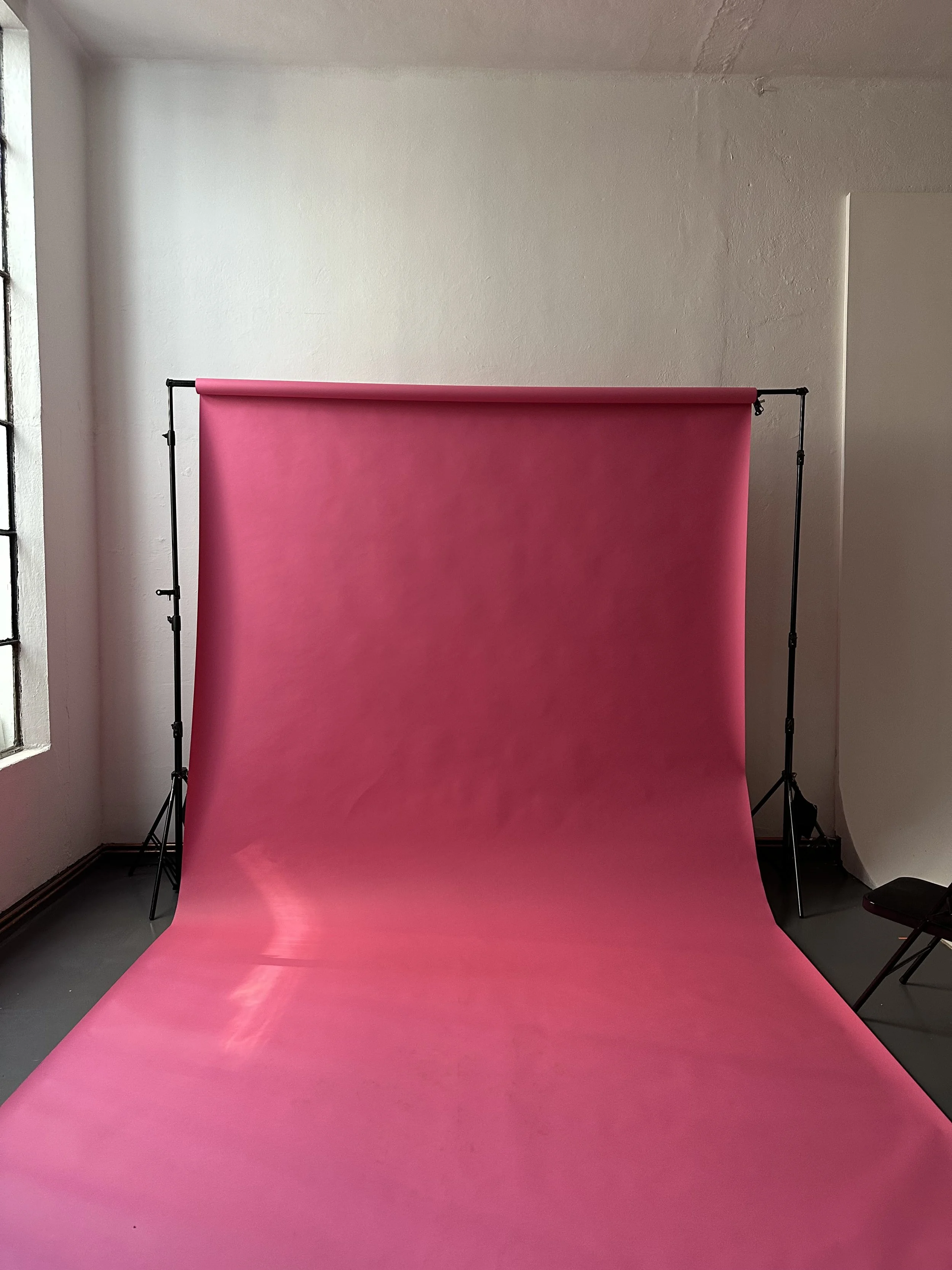 Uno sfondo di studio fotografico con un fondale rosa e un supporto per fondale, in una stanza con pareti bianche e una finestra a sinistra.