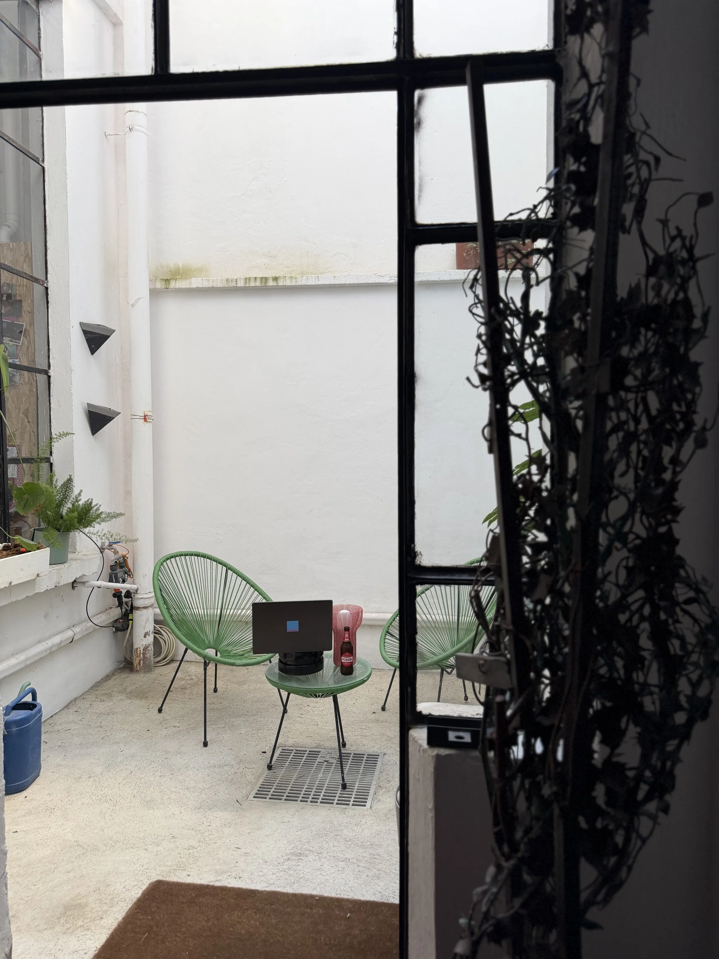 Un cortile interno con due sedie di plastica verdi, un tavolino con un computer portatile, una bottiglia di birra e una lampada rossa, e alcune piante. L'immagine è vista attraverso una porta semicatturata.