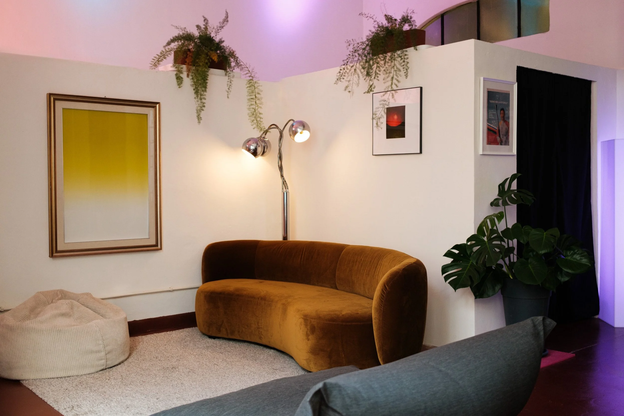 Soggiorno con divano arancione, cuscino beige, lampada da terra con due luci, quadro e piante sopra una parete bianca, con luci soffuse e luci ambientali colorate.