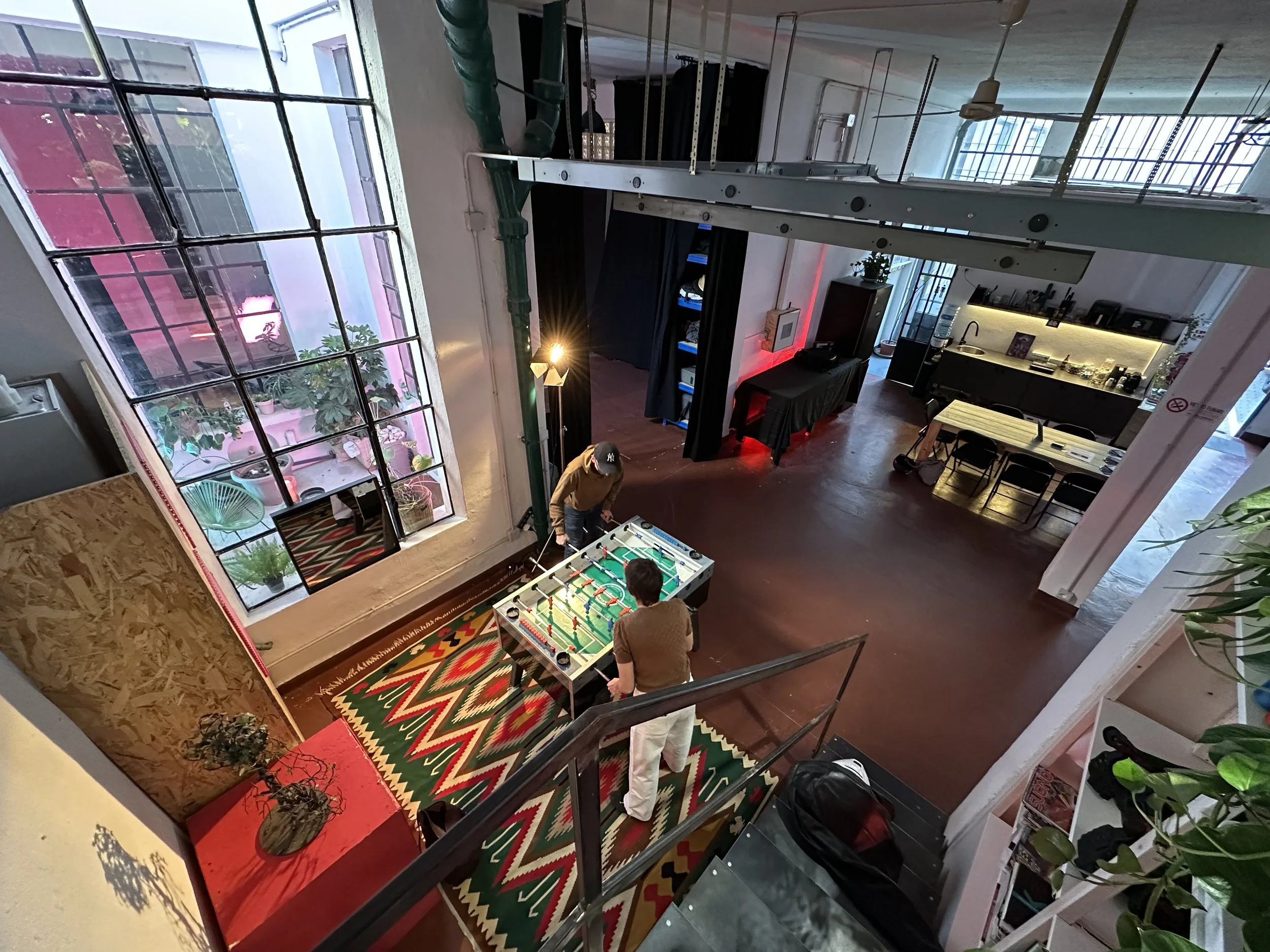 Interno di un loft con una zona cucina moderna con illuminazione a LED, una sala con un tappeto colorato e un tavolo da calcio balilla, piante in vaso, grandi finestre con vista sulla città e alcune persone.