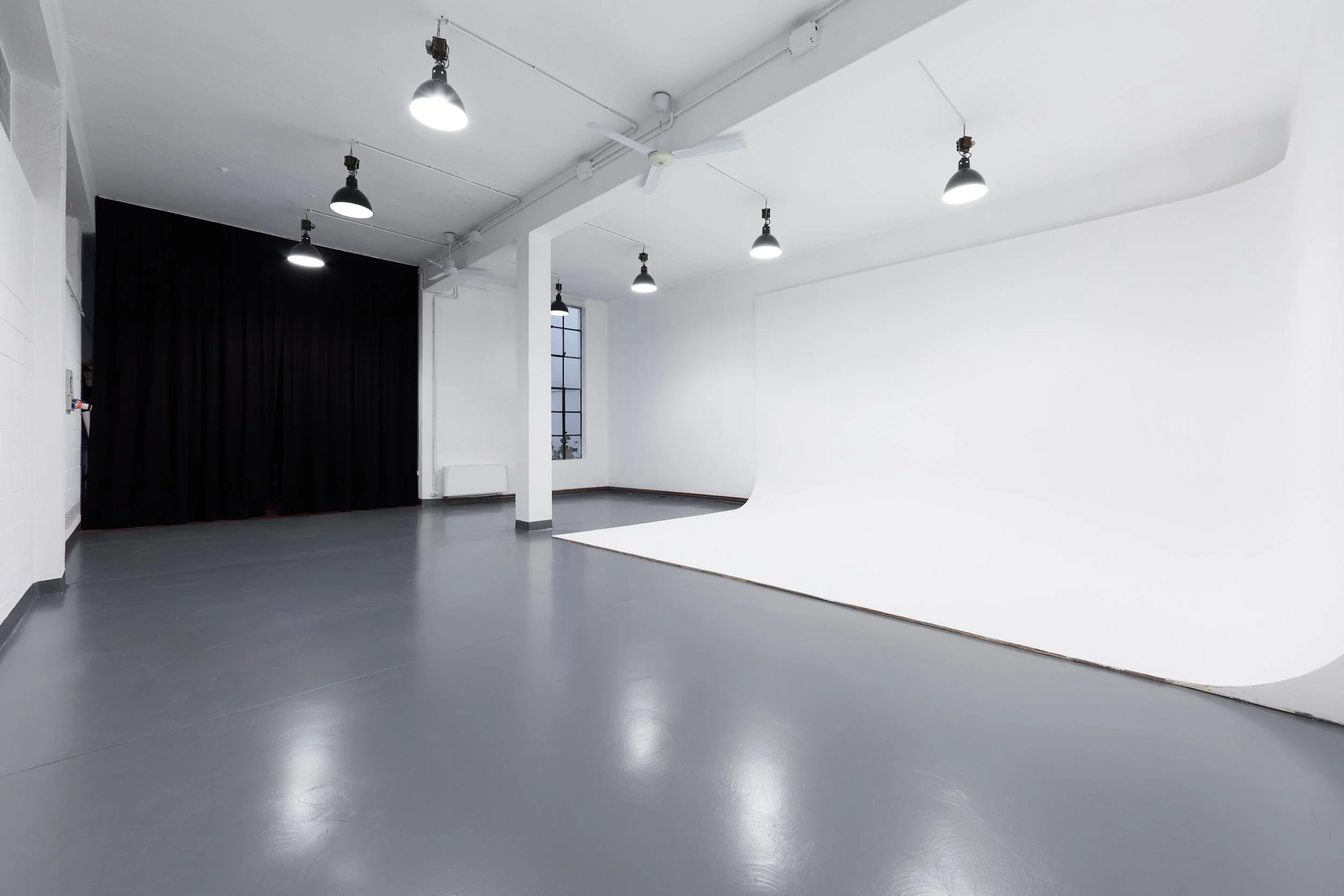 Studio fotografico vuoto con parete bianca curva, pavimento grigio, tende nere, finestra e luci da studio.