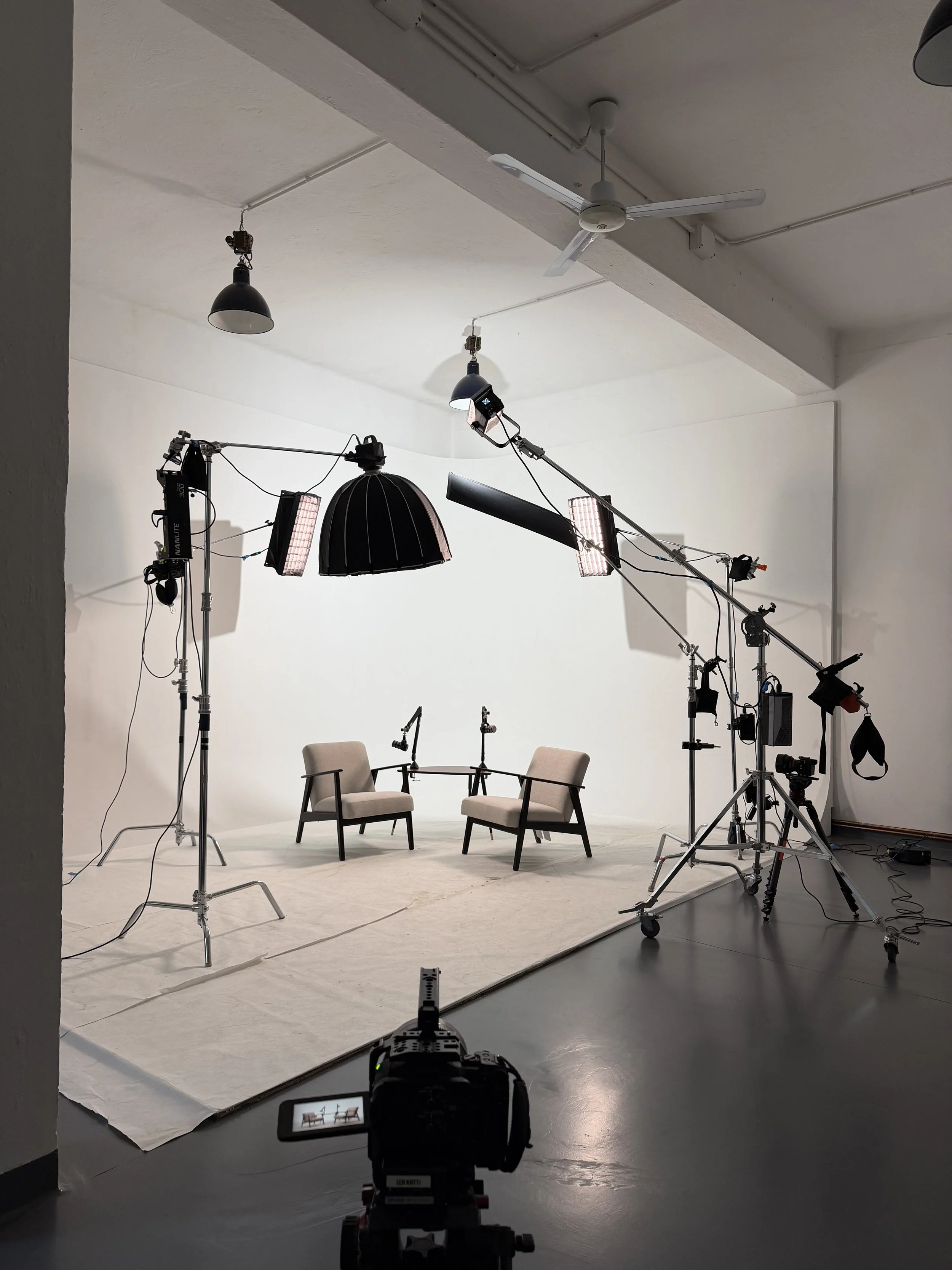 Set di luci e attrezzature per una sessione fotografica o di registrazione. Sullo sfondo, due sedie e un tavolo con due microfoni.
