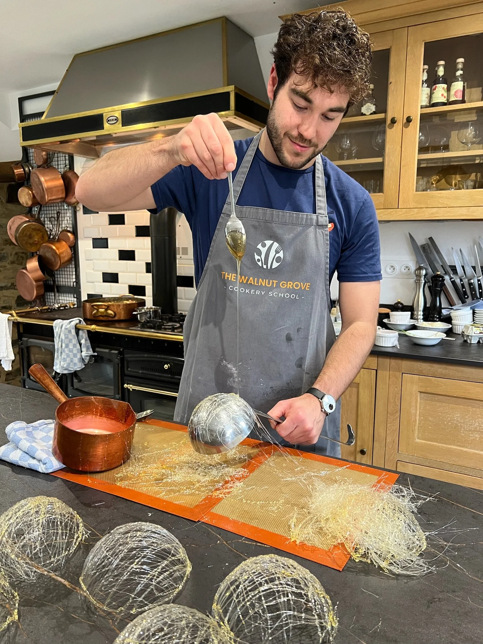 A man in a gray apron with the logo for The Walnut Grove Cookery School drizzles caramel over a sphere-shaped dessert on a silicone mat in a cozy kitchen, with multiple copper pots hanging on the wall and various bowls and utensils on the countertop.