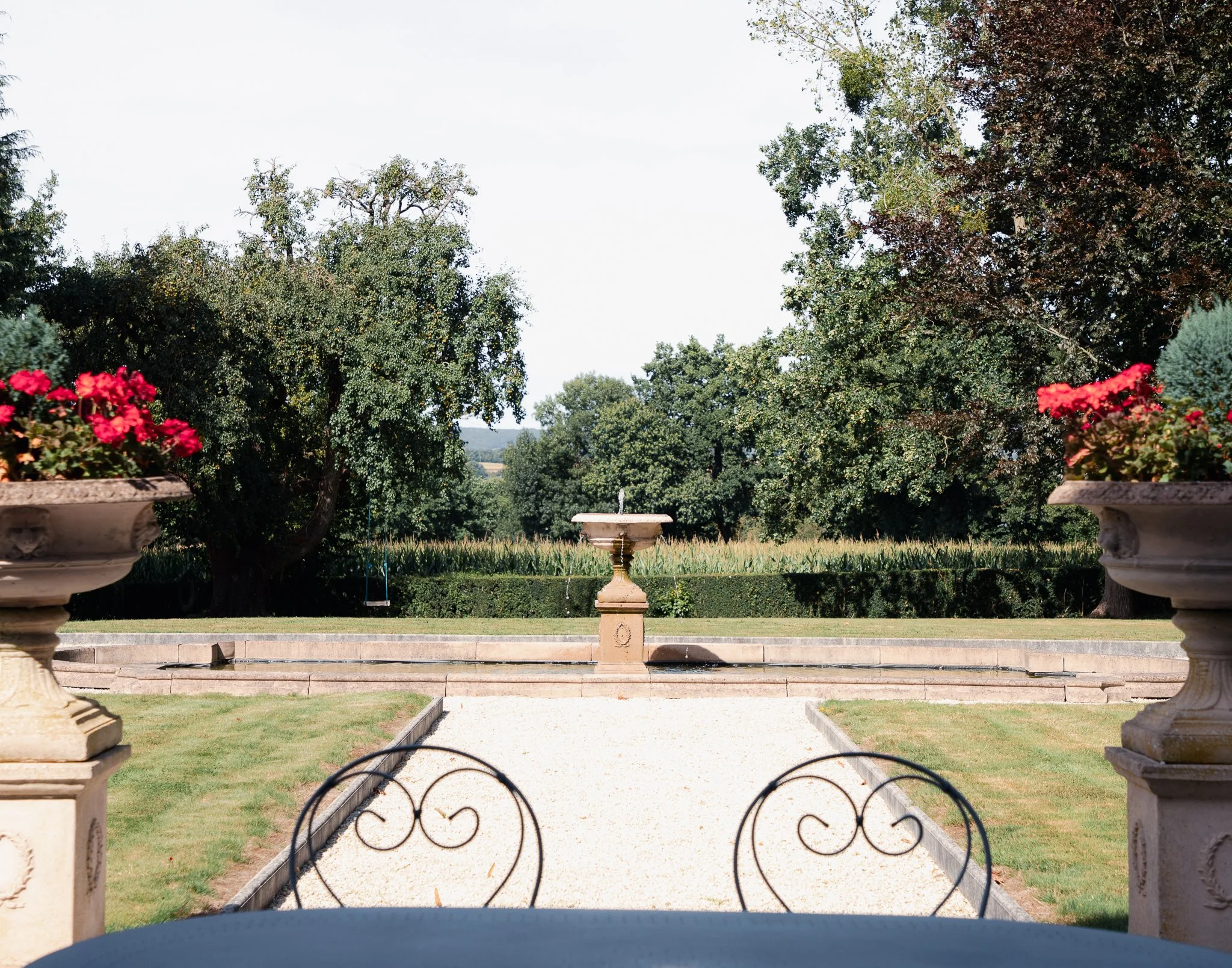 View of a garden with a stone fountain at the center, flanked by large stone planters with red flowers, with trees and a clear sky in the background.