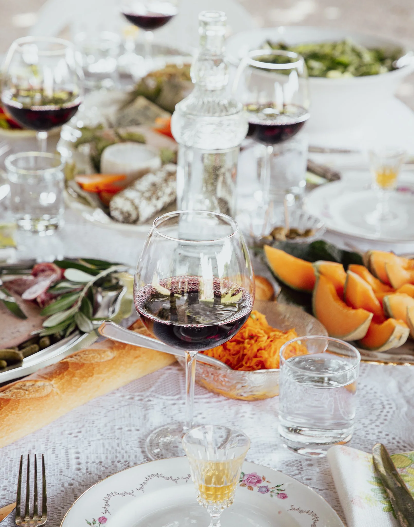 A table set for a meal with glasses of red wine, water, a loaf of bread, sliced cantaloupe, a dish of grated carrots, a plate of meat, a bowl of salad, and various dishes and utensils.