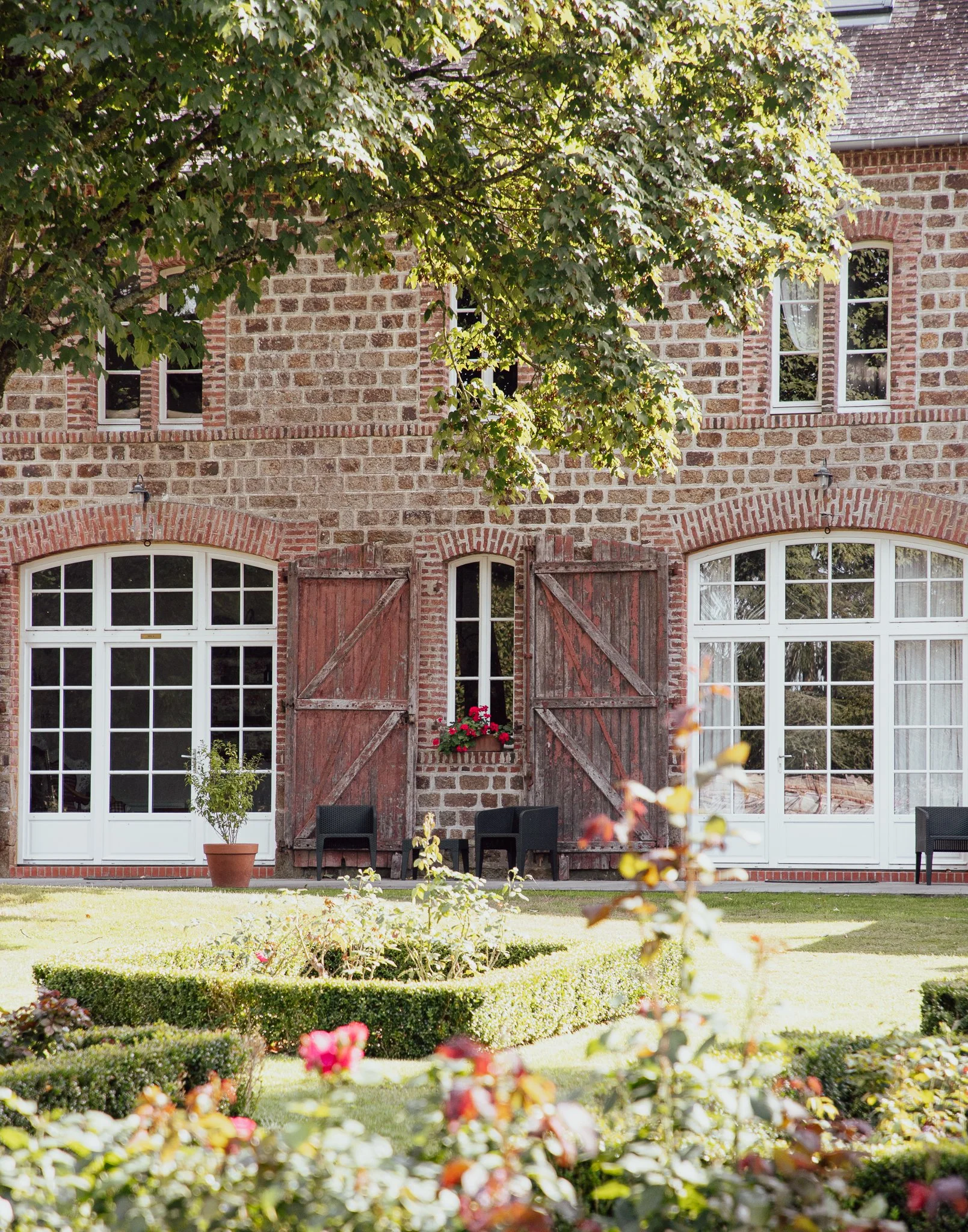 Brick house with large windows, open wooden shutters, potted plants, outdoor seating, and a manicured garden with flower beds and hedge borders.