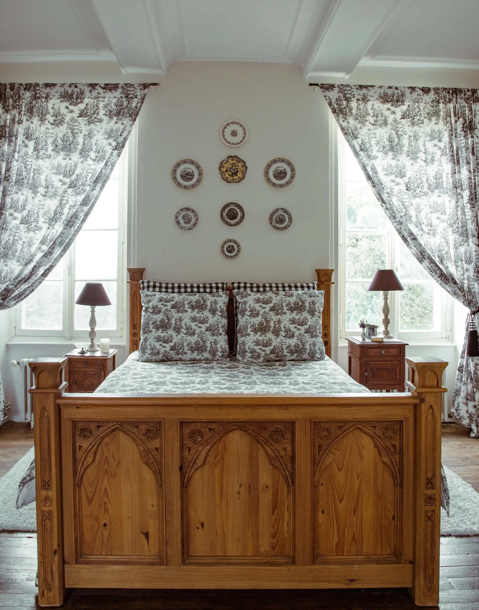 A bedroom with a wooden bed frame and matching nightstands, floral patterned bedding, checkered pillows, and windows with patterned curtains. Decorative plates are hung on the wall above the bed.