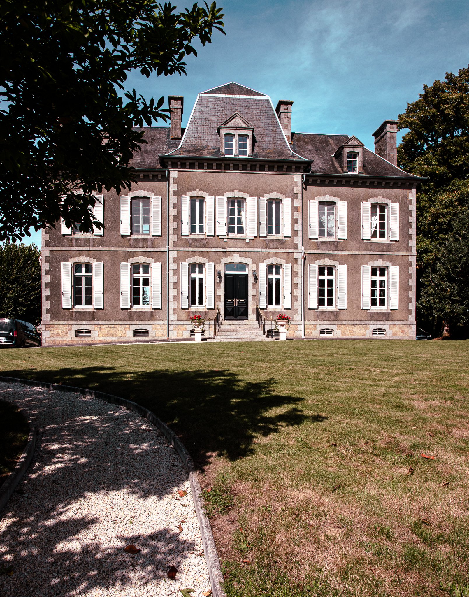 A large, historic mansion with gray stone walls and white shutters, surrounded by trees and a well-maintained lawn, with a gravel pathway on the left side.