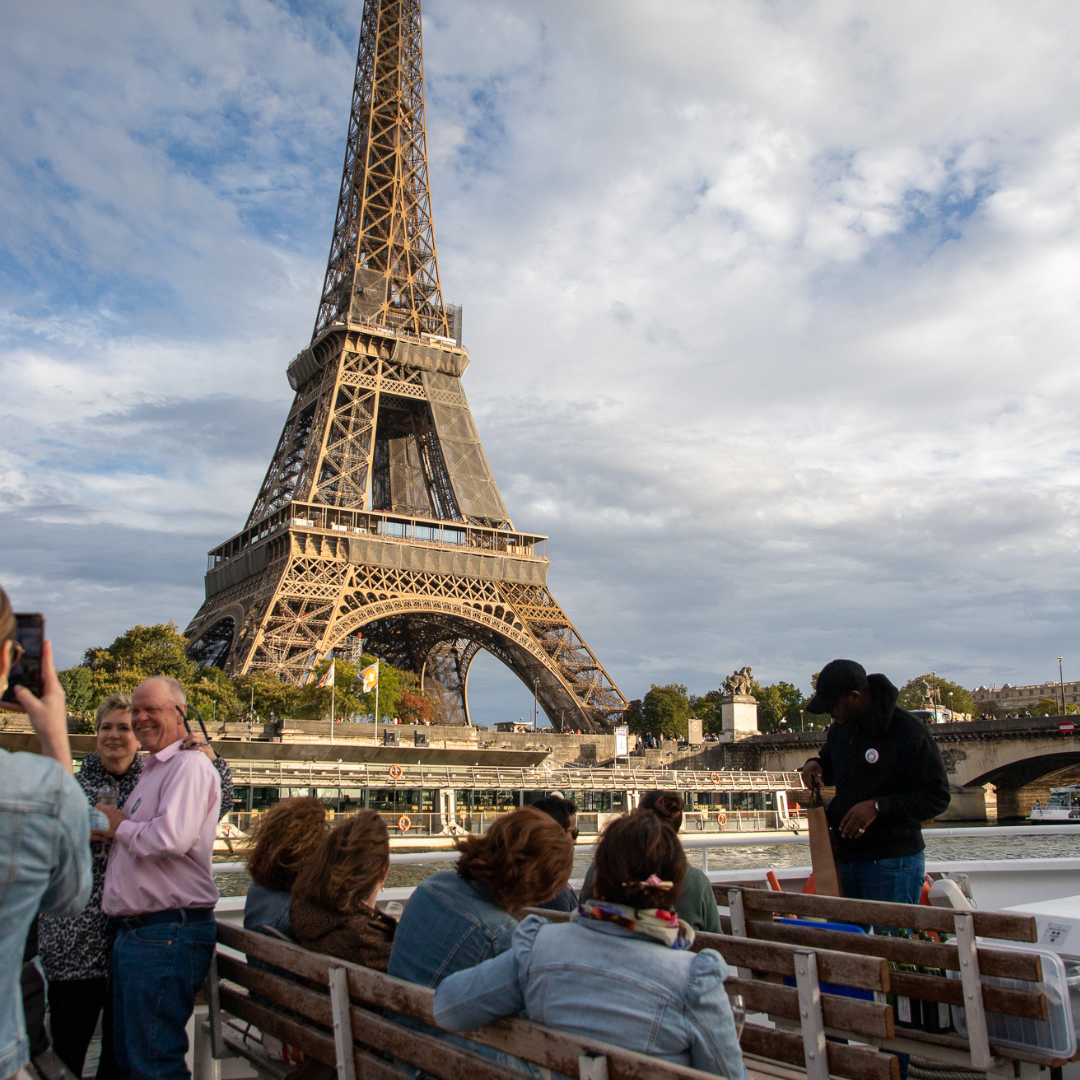 People sitting and standing on a boat tour next to the Seine River with the Eiffel Tower in Paris, France, during daytime.