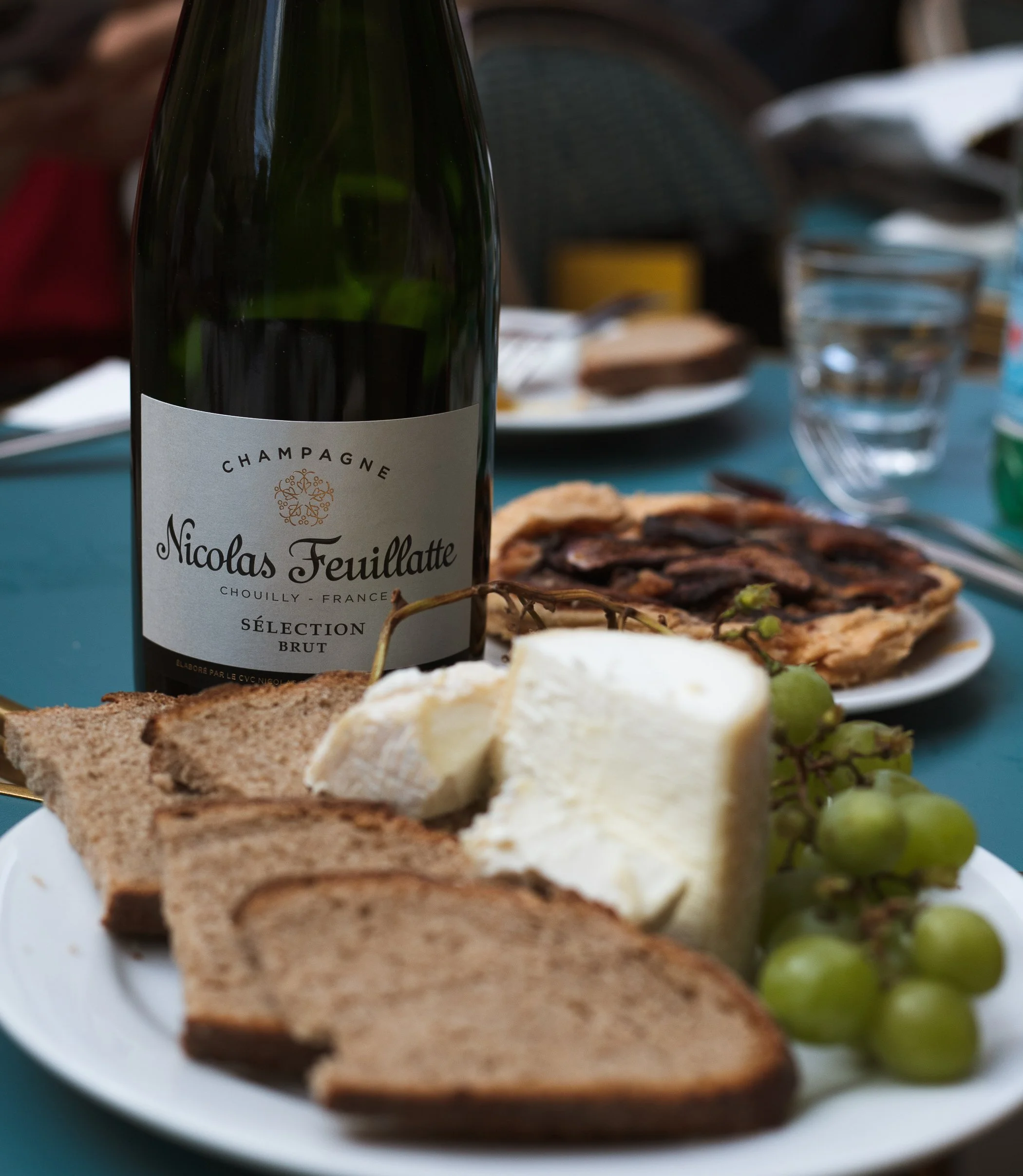 Plate with bread, cheese, grapes, and a bottle of Nicolas Feuillatte champagne, with a slice of pizza and glasses of water in the background.