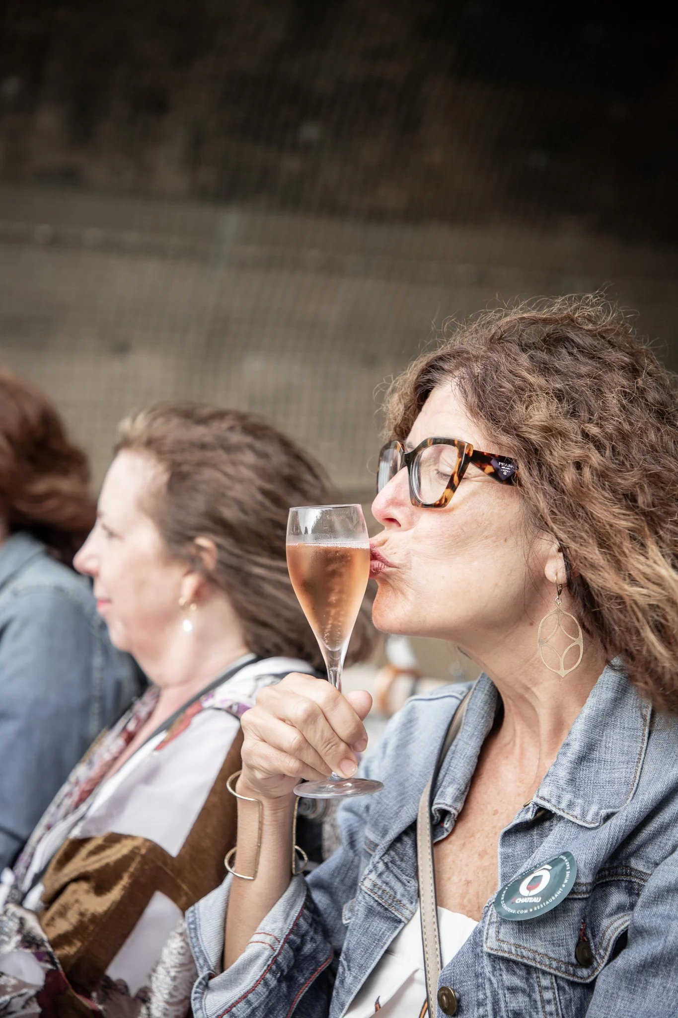 A woman with curly hair, glasses, and earrings is holding a glass of rosé wine or champagne and kissing it. She is sitting next to a woman with straight hair and wearing a multi-colored patterned scarf or shawl.
