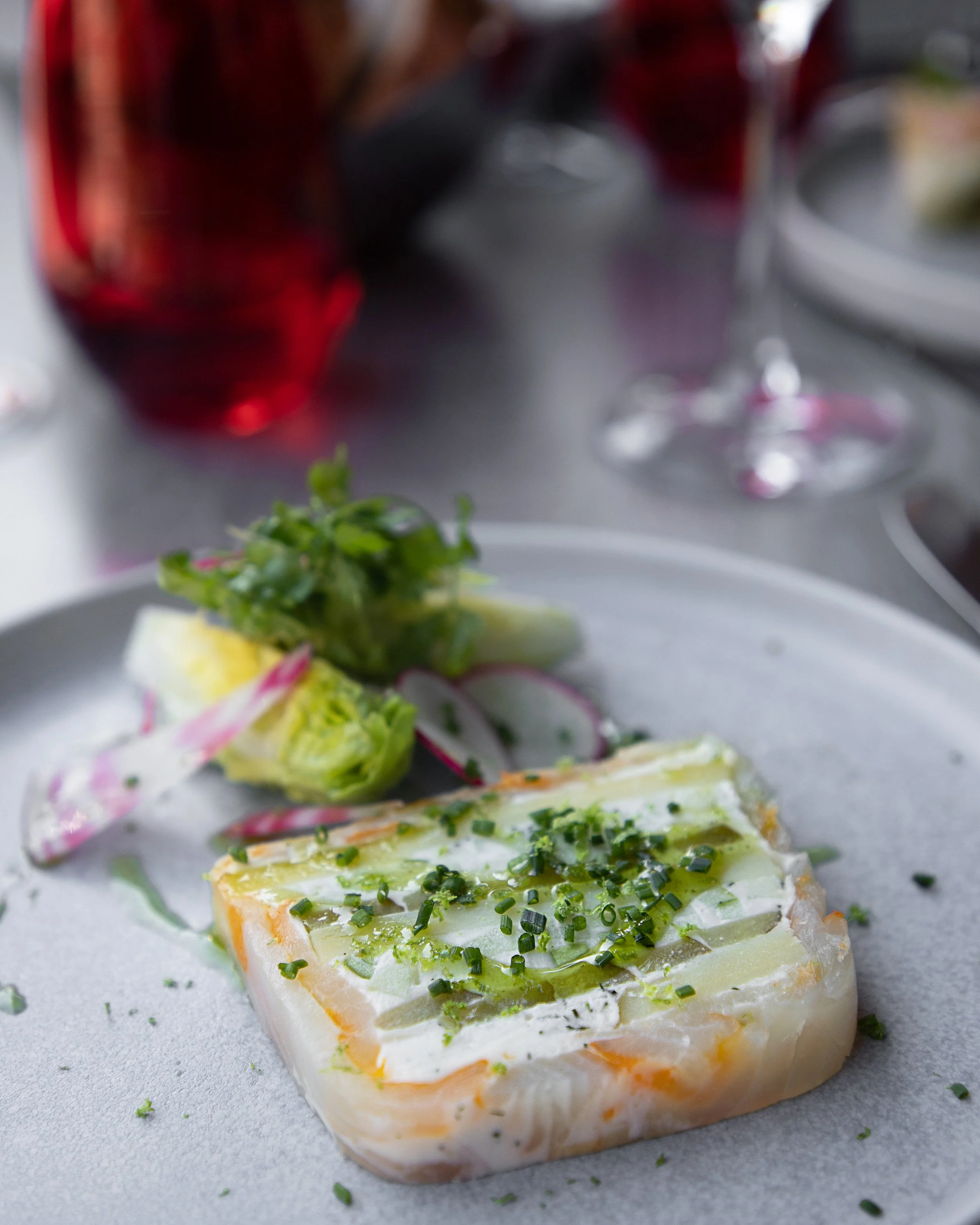 A plate with sliced vegetable terrine topped with chopped chives, served with a side salad of lettuce and radish slices, on a gray plate in a restaurant setting with a red beverage and a wine glass in the background.