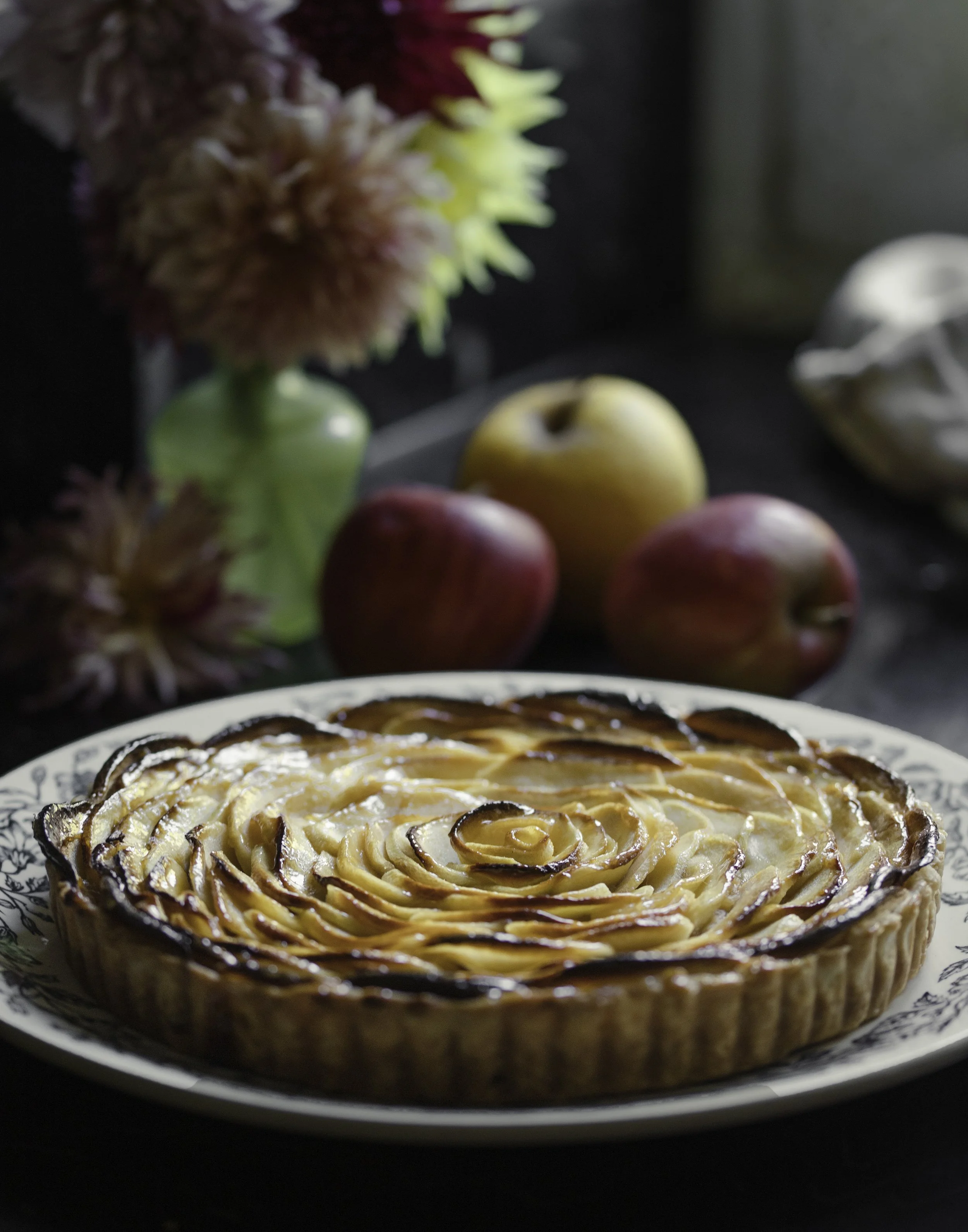 A baked apple tart on a decorative plate with a floral pattern, in front of a dark background with three apples and a bouquet of flowers in a green vase.