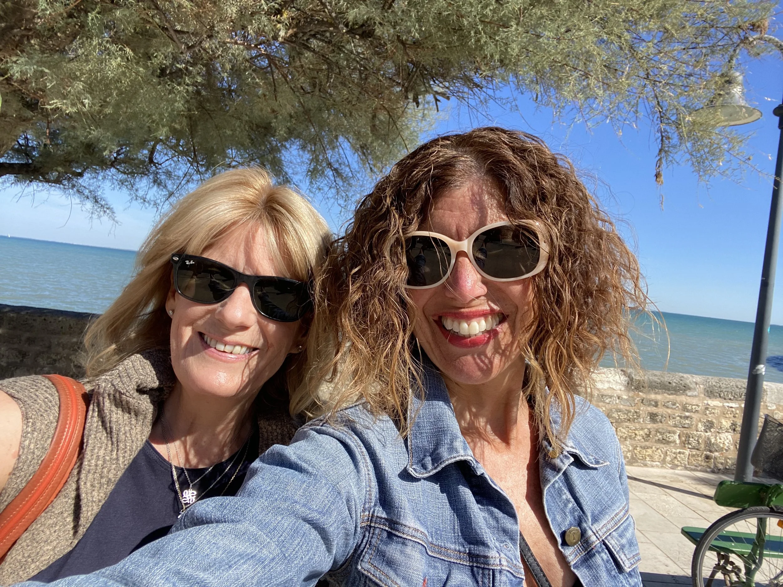 Two women smiling for a selfie at the beach, with ocean, trees, and a bicycle in the background.