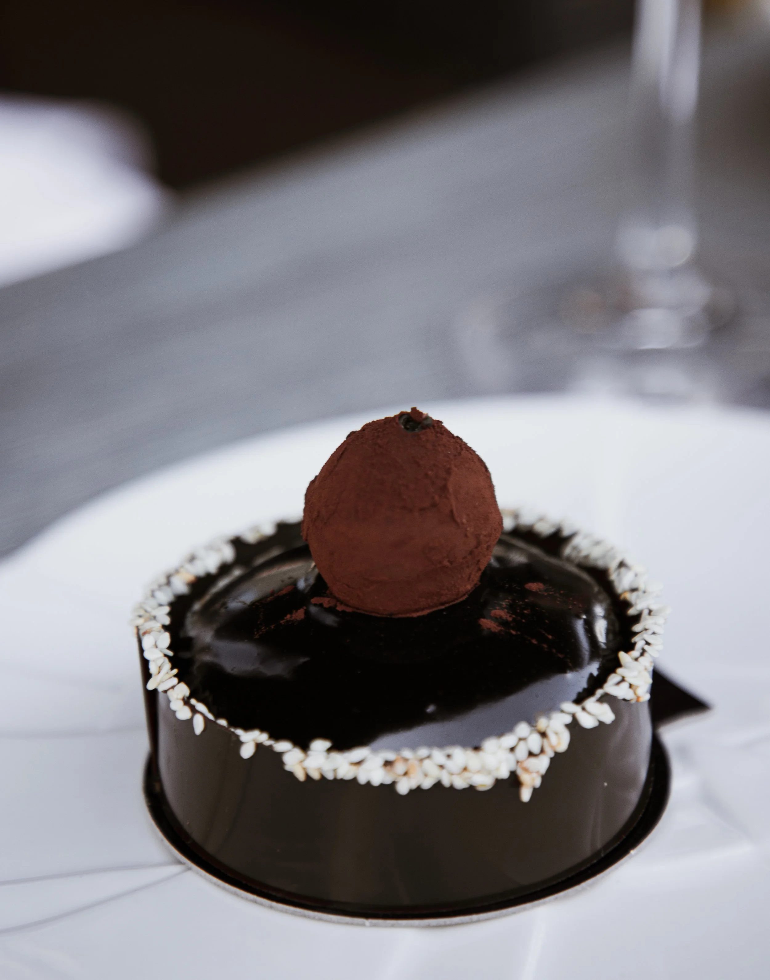 Chocolate mousse cake topped with a cocoa-dusted truffle, served on a white plate with a decorative edge.