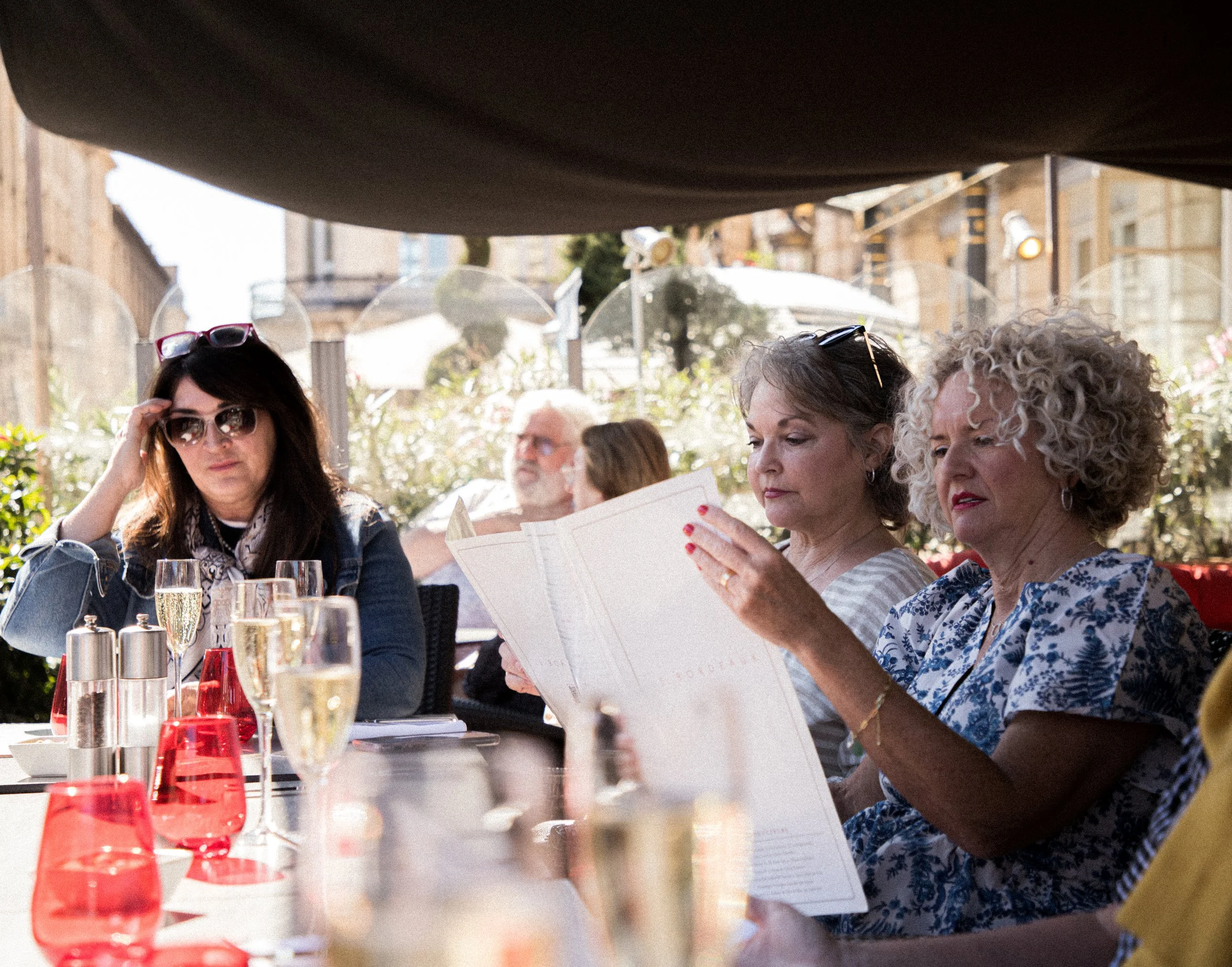Group of women and men dining outdoors at a restaurant, with champagne glasses on the table, some women reading menus, in a sunny urban setting with buildings and greenery in the background.