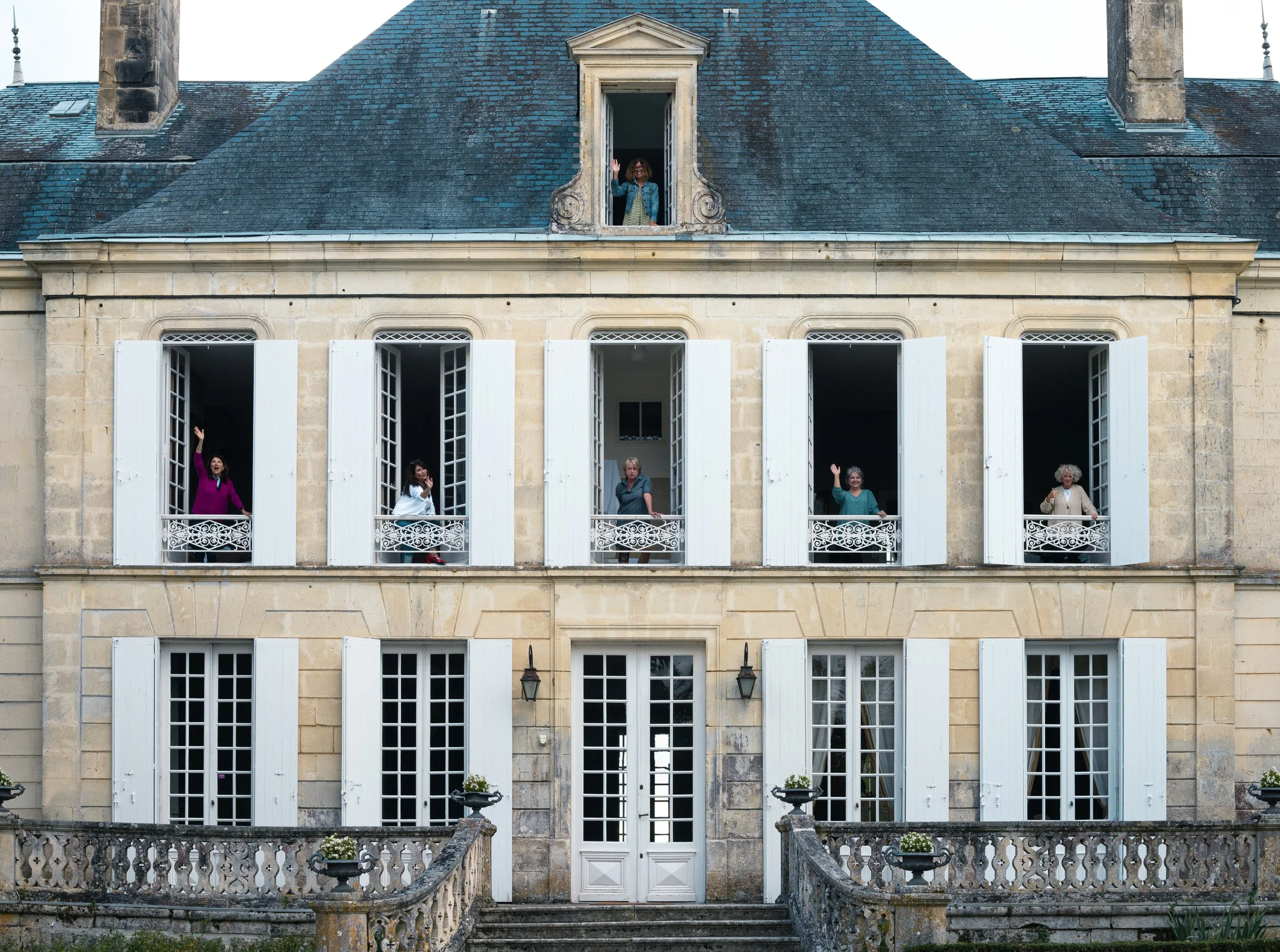 An elegant three-story building with white shutters open on all windows and a person waving from a window on the top floor. The facade is beige stone with a central entrance. Several women are seen talking and waving from the windows.