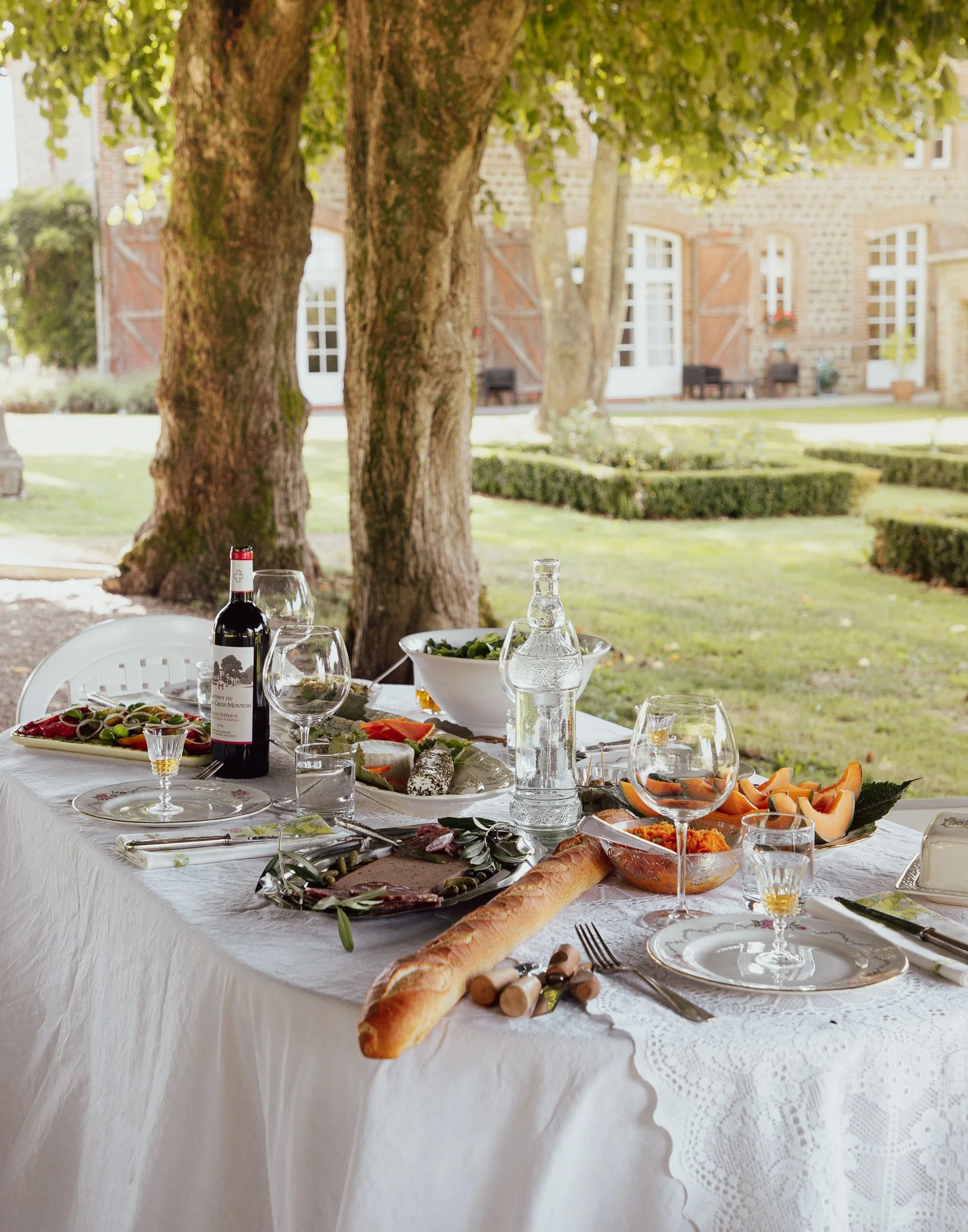 Outdoor dining table with bread, wine, glasses, salad, and other dishes set under a large tree on a sunny day.