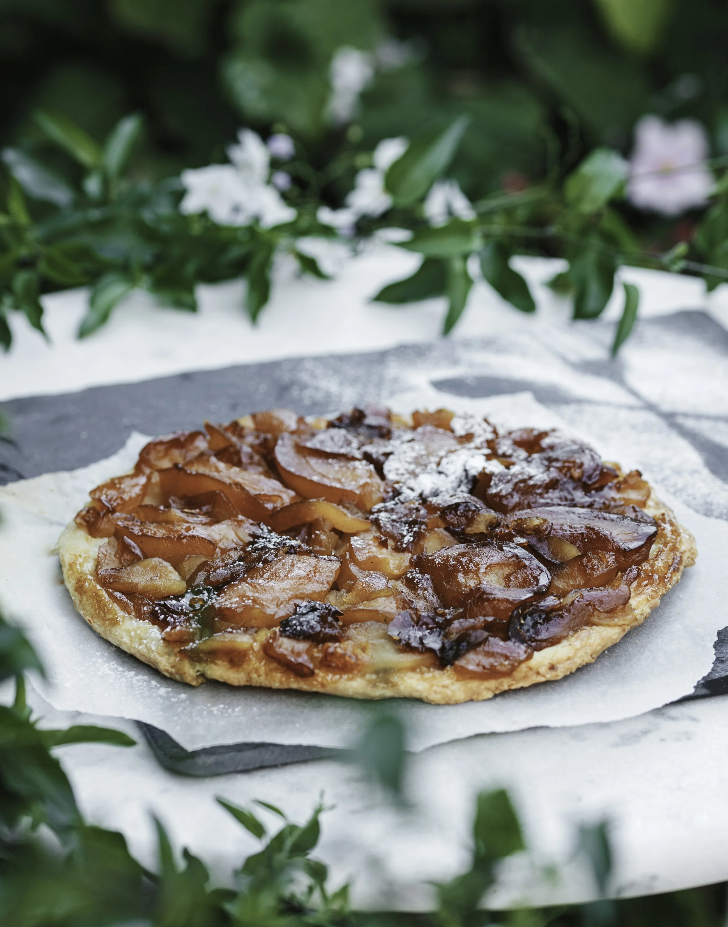 Fresh peach and cherry tart on a black slate serving board, dusted with powdered sugar, outdoors with green foliage in the background.