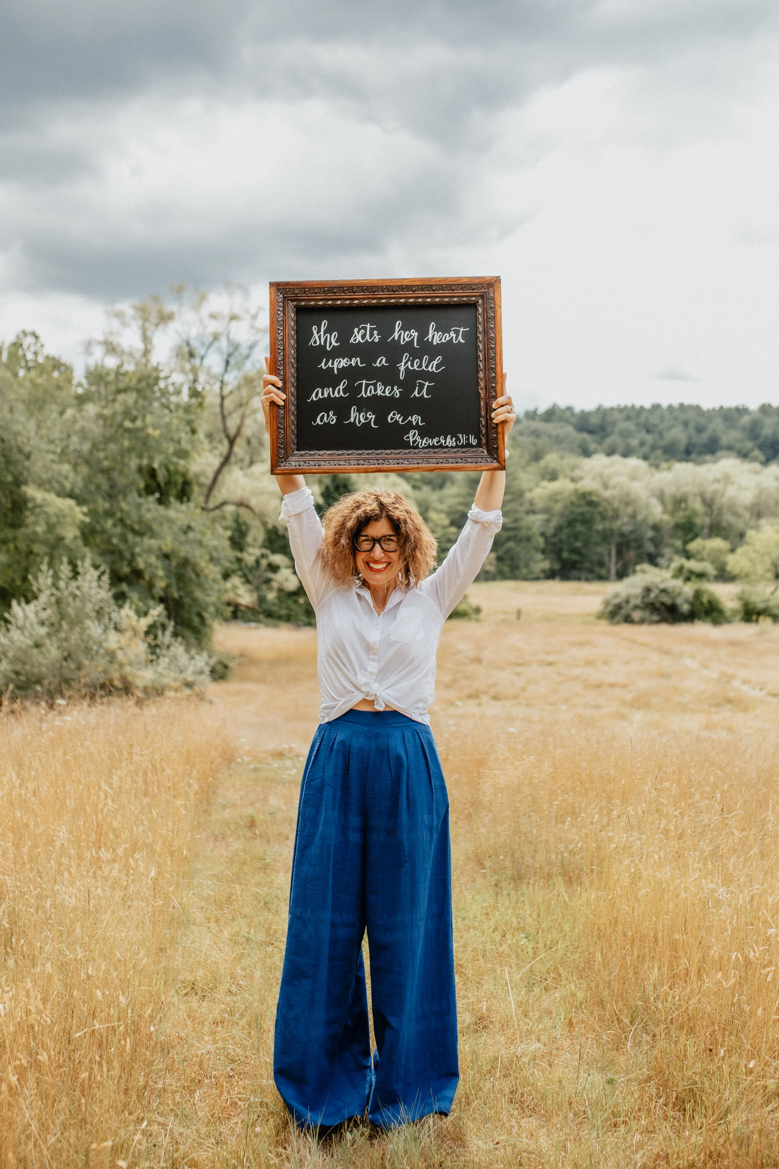 Woman with curly hair and glasses holding a framed blackboard with a Bible quote in an open field with trees, under a cloudy sky.