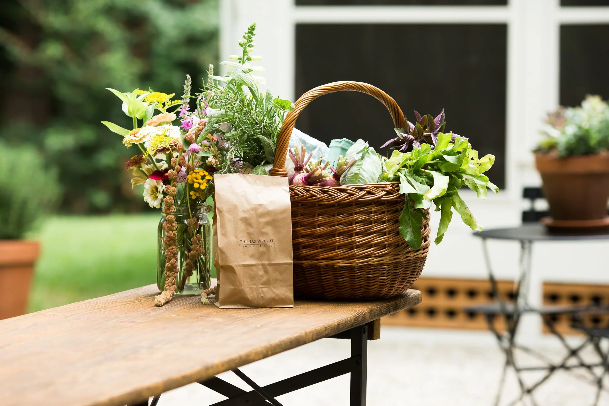A wooden table holding a wicker basket and a glass jar filled with fresh flowers, with a paper bag labeled 'Phineas Wright' placed next to the basket, outdoors with blurred greenery and potted plants in the background.