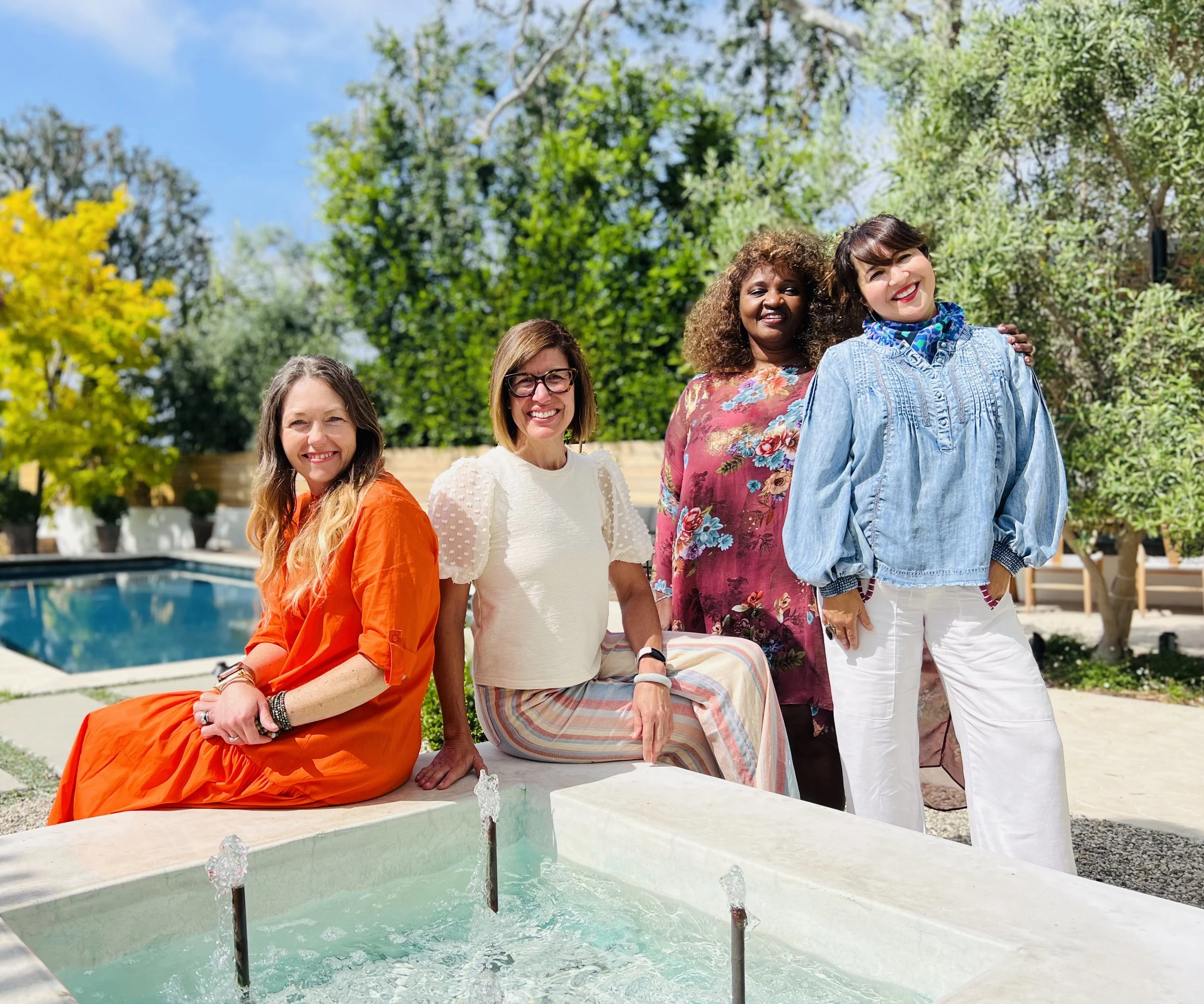 Four women sitting and standing outdoors in a garden with trees, a swimming pool, and a fountain in the foreground under a blue sky.