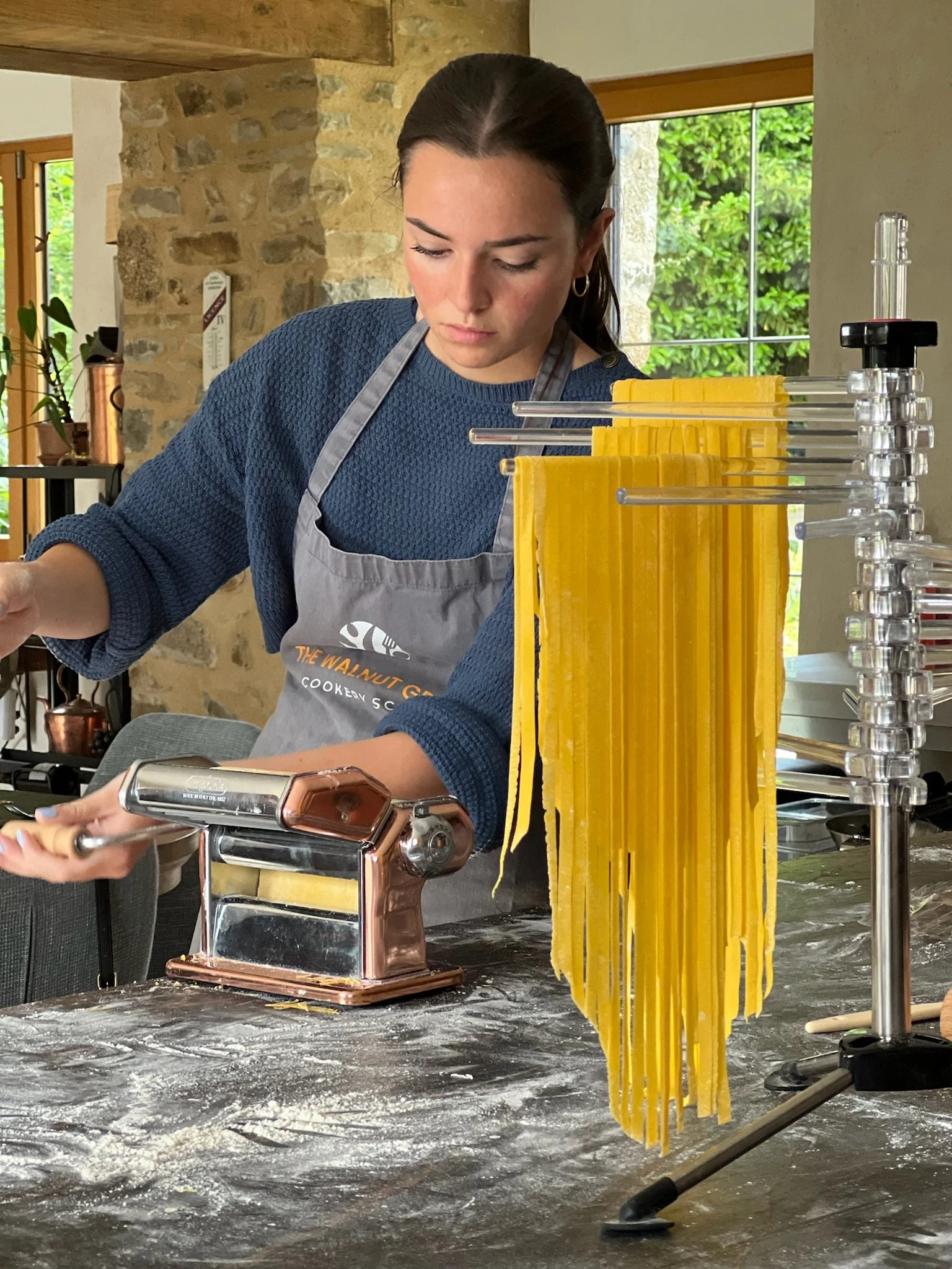 A woman in a gray apron with a logo, standing in a kitchen, making fresh pasta using a pasta machine. Dried pasta strands hang on a rack nearby, and the kitchen has rustic stone walls and large windows showing greenery outside.