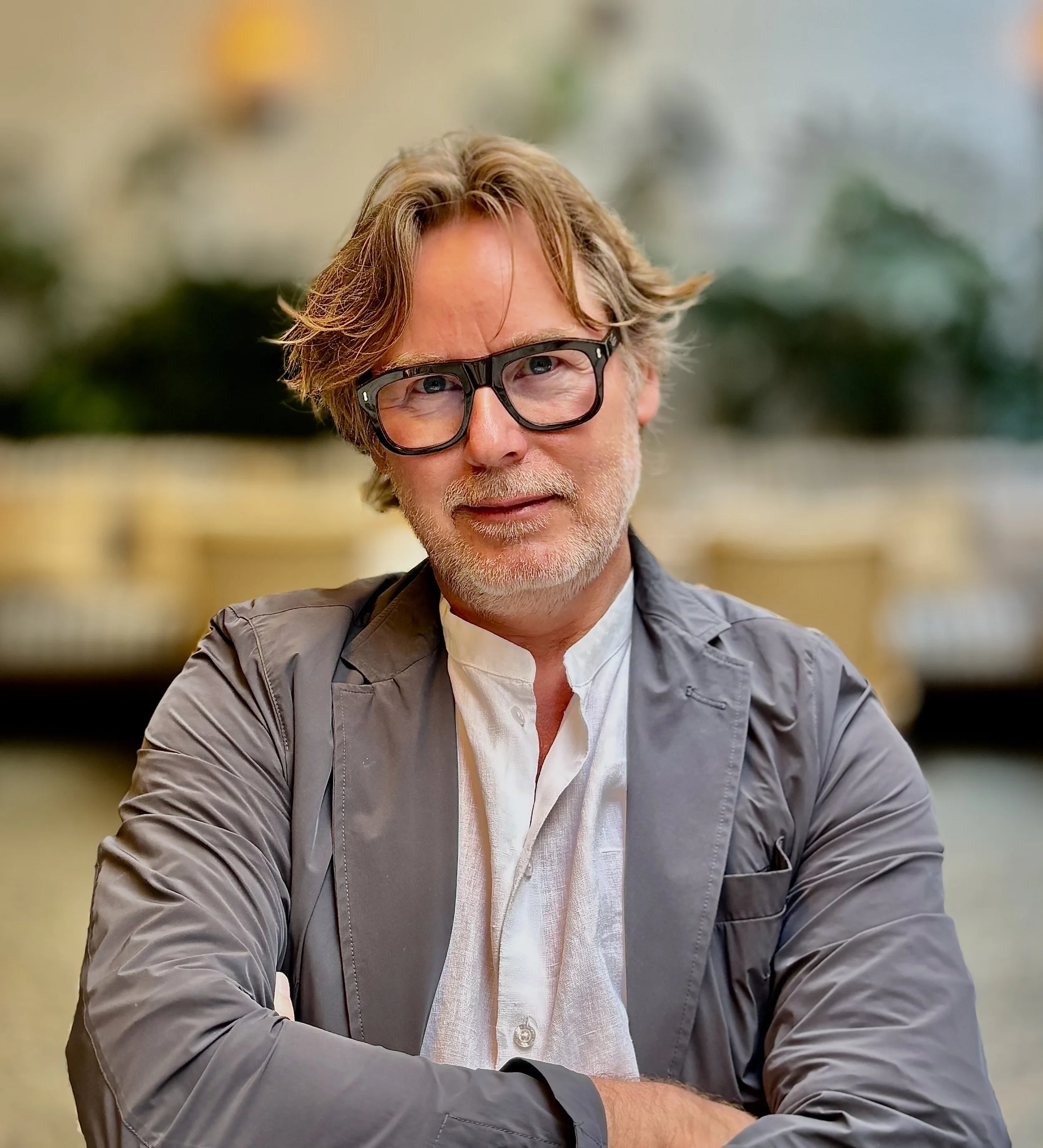 En mand med skæg og briller smiler, iført en hvid skjorte og en grå jakke, med en blurred baggrund.