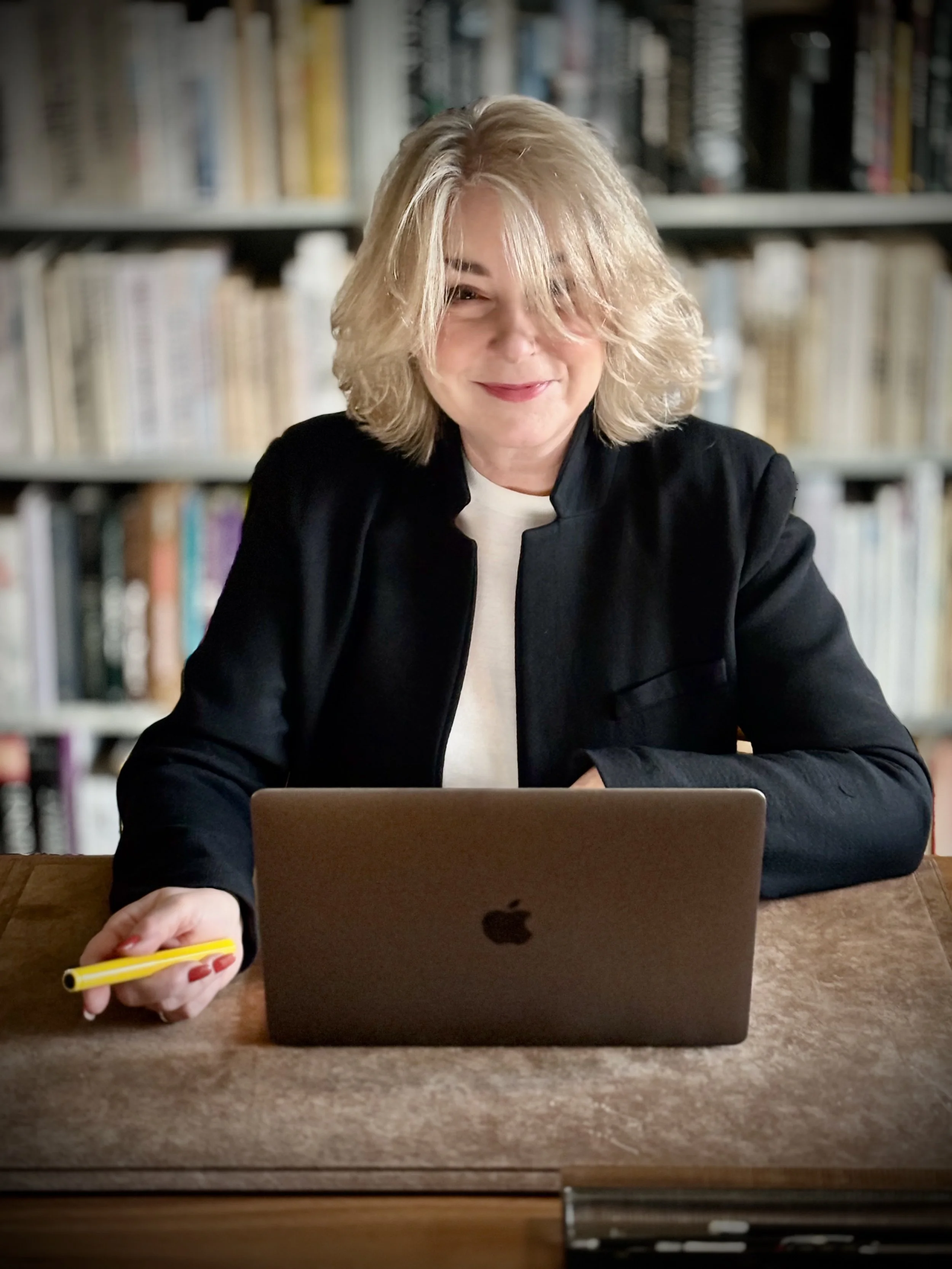 En kvinde med blond, bølget hår sidder ved et skrivebord foran en åben MacBook, holder en gul marker. Bag hende er der en boghylde fyldt med bøger.