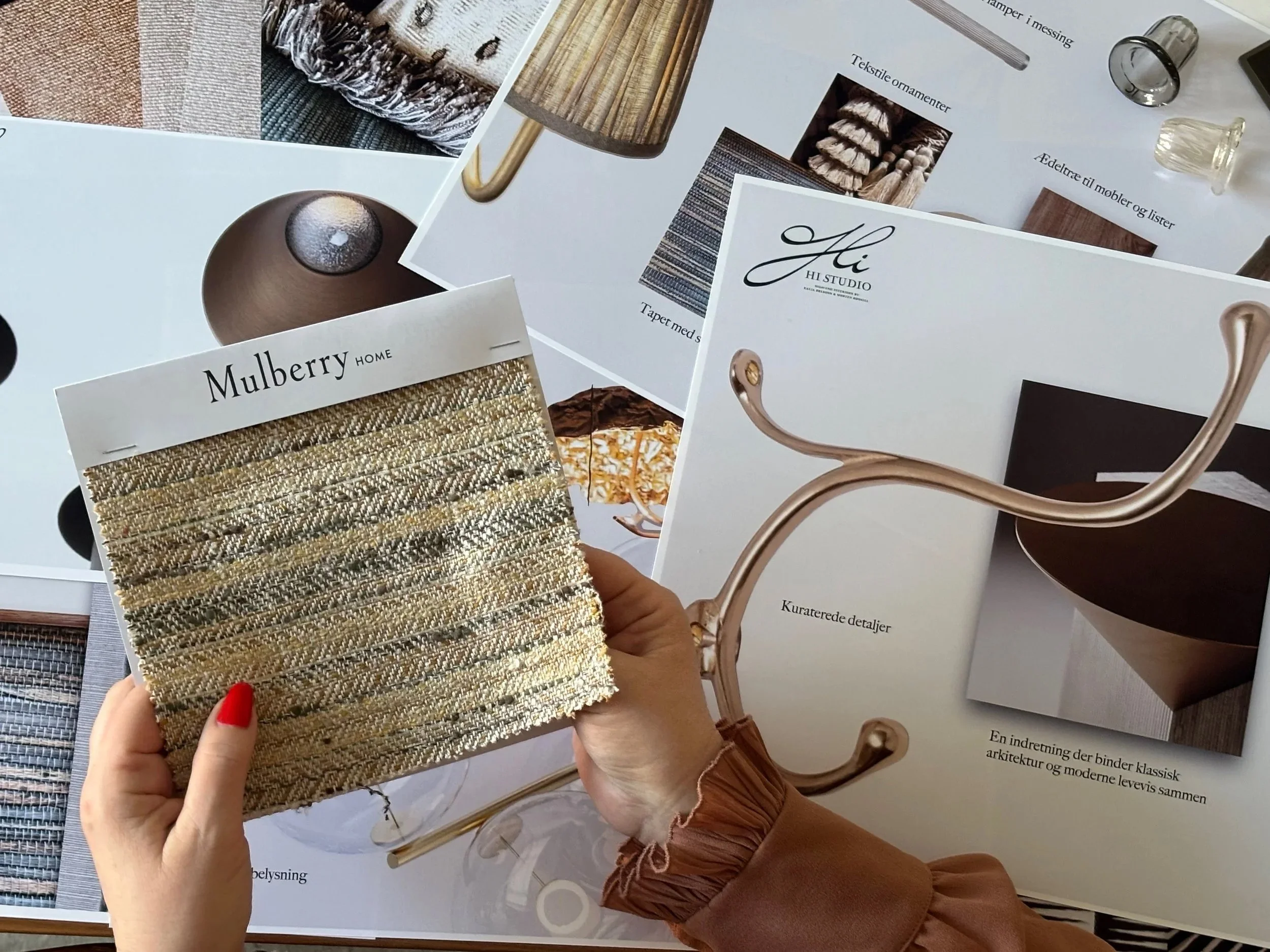 Person holder vægprøve af tekstil med guld- og beige nuancer foran åbne brochurer om indretning og design.