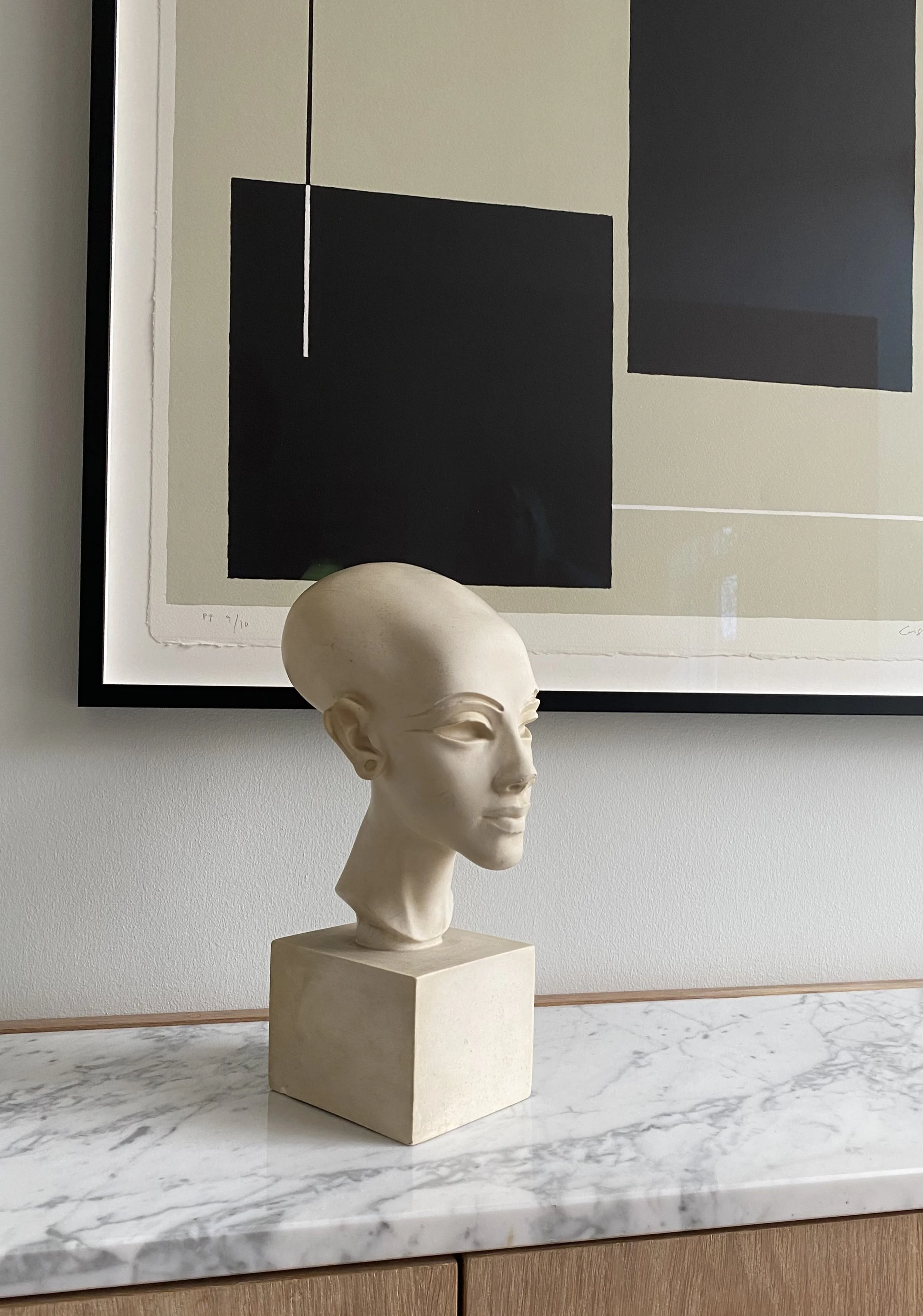 En skulptur af en kvindelig figur med kort hår, placeret på en beige firkantet base på et marmorbord, foran et abstrakt kunstværk med sorte, grå og beige farver.