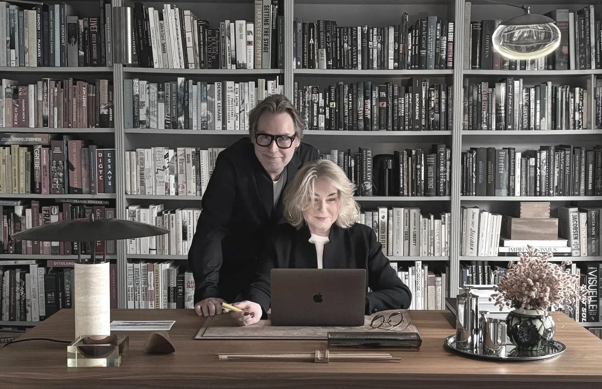 To personer i et kontor med bogreoler bag dem. En mand står bag en kvinde, der sidder foran en laptop på et skrivebord. Der er en plante, en lampe, notesbog, briller og kopper på skrivebordet.