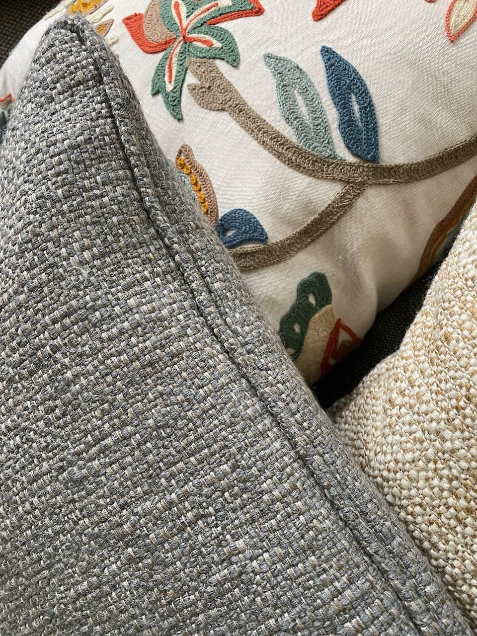 En pude med broderet blomster- og bladmotiv i nuancer af rød, grøn, blå og beige, omgivet af tekstiler med forskellige vævninger.