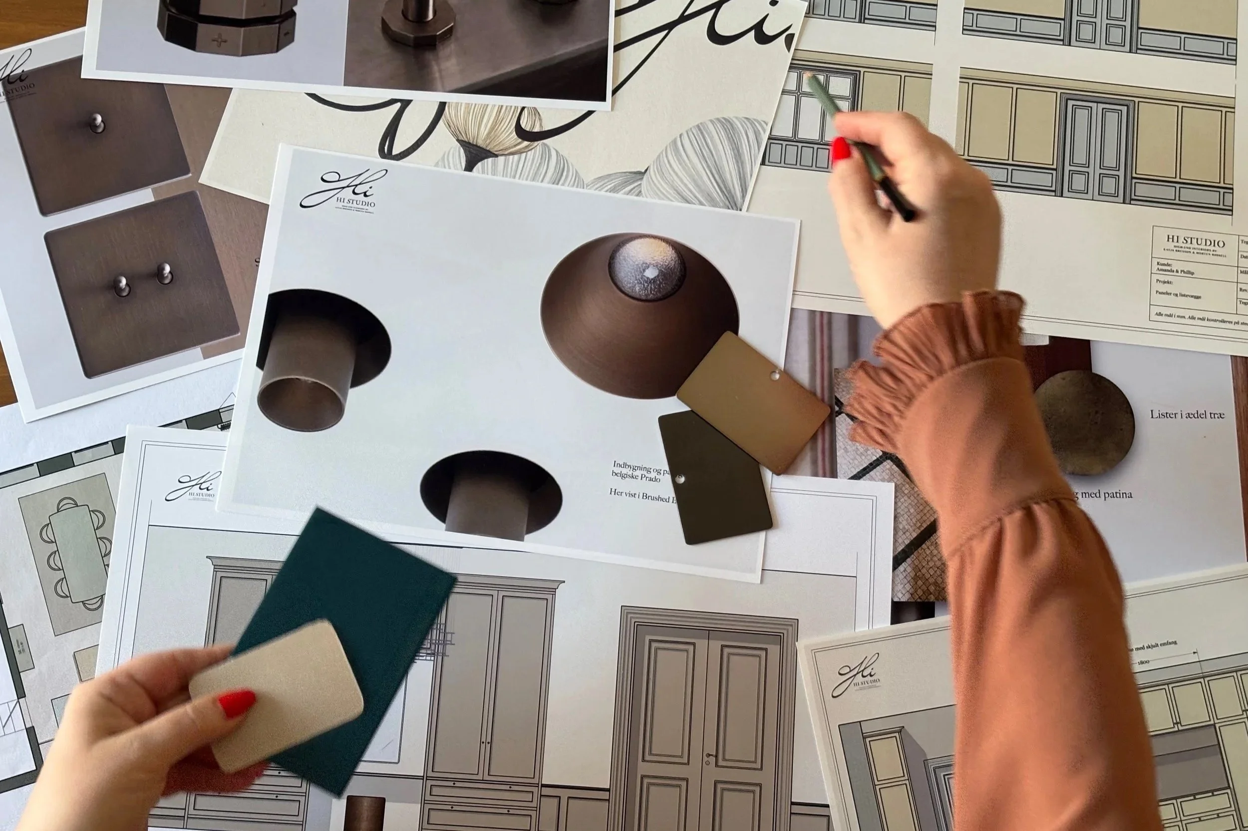 Billede af en hånd med rød negl, der holder farveprøver, mens en anden hånd skriver på ark med indretningstegninger og farvemønstre. Der er flere ark med designskitser og farvematerialer spredt ud på et bord.