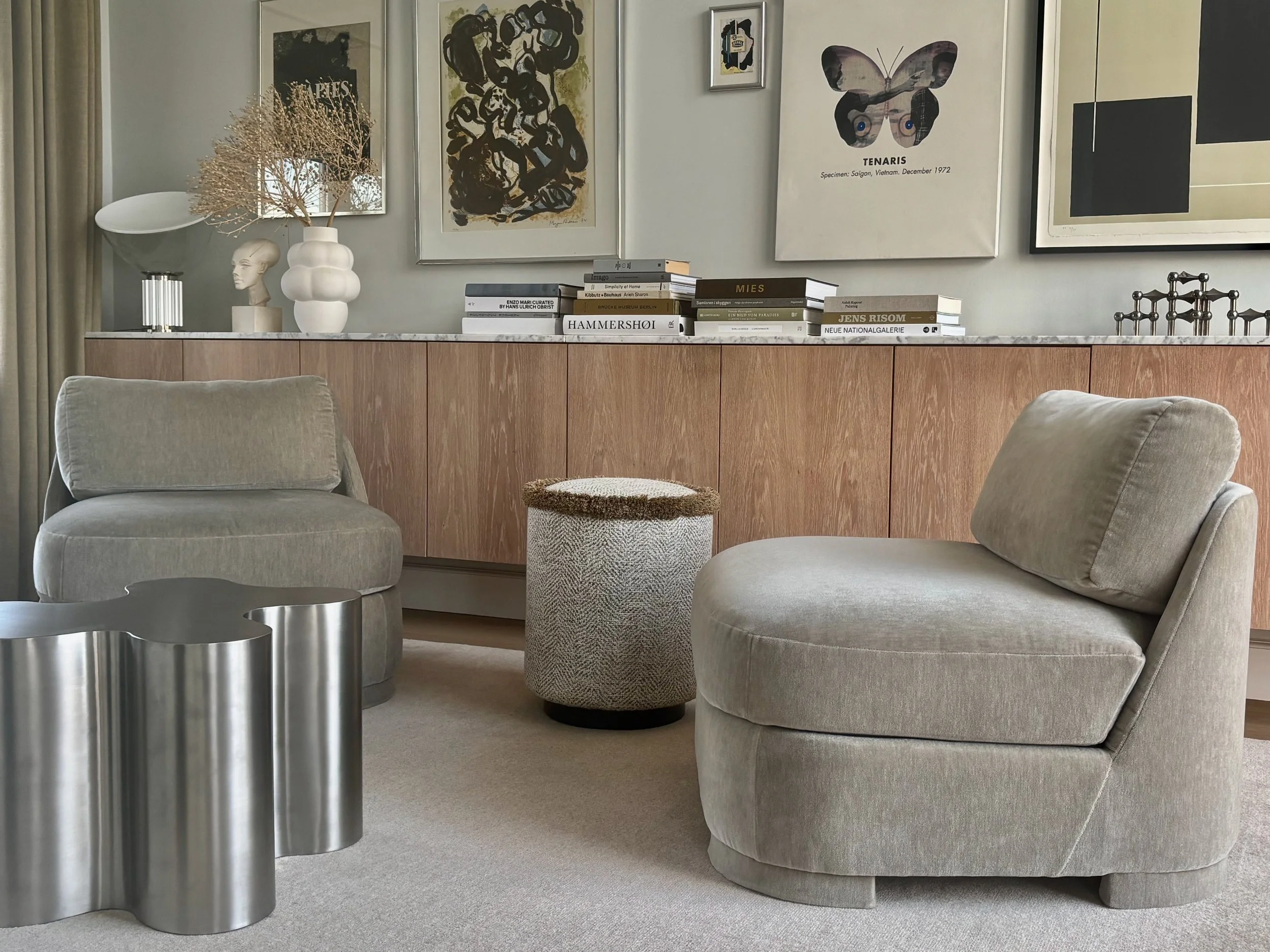 Et moderne stueindretning med to beige-velourpolstrede loungestole, en rund beige uldtæppe, en metallisk skulptur i form af tre forbundne cylindere, en beige stofpuf med brunt kanvaskant, en trælavet mediebuffet med forskellige kunstværker og bøger på toppen, samt vægkunst inklusive en butterfly-plakat og abstrakte billeder.